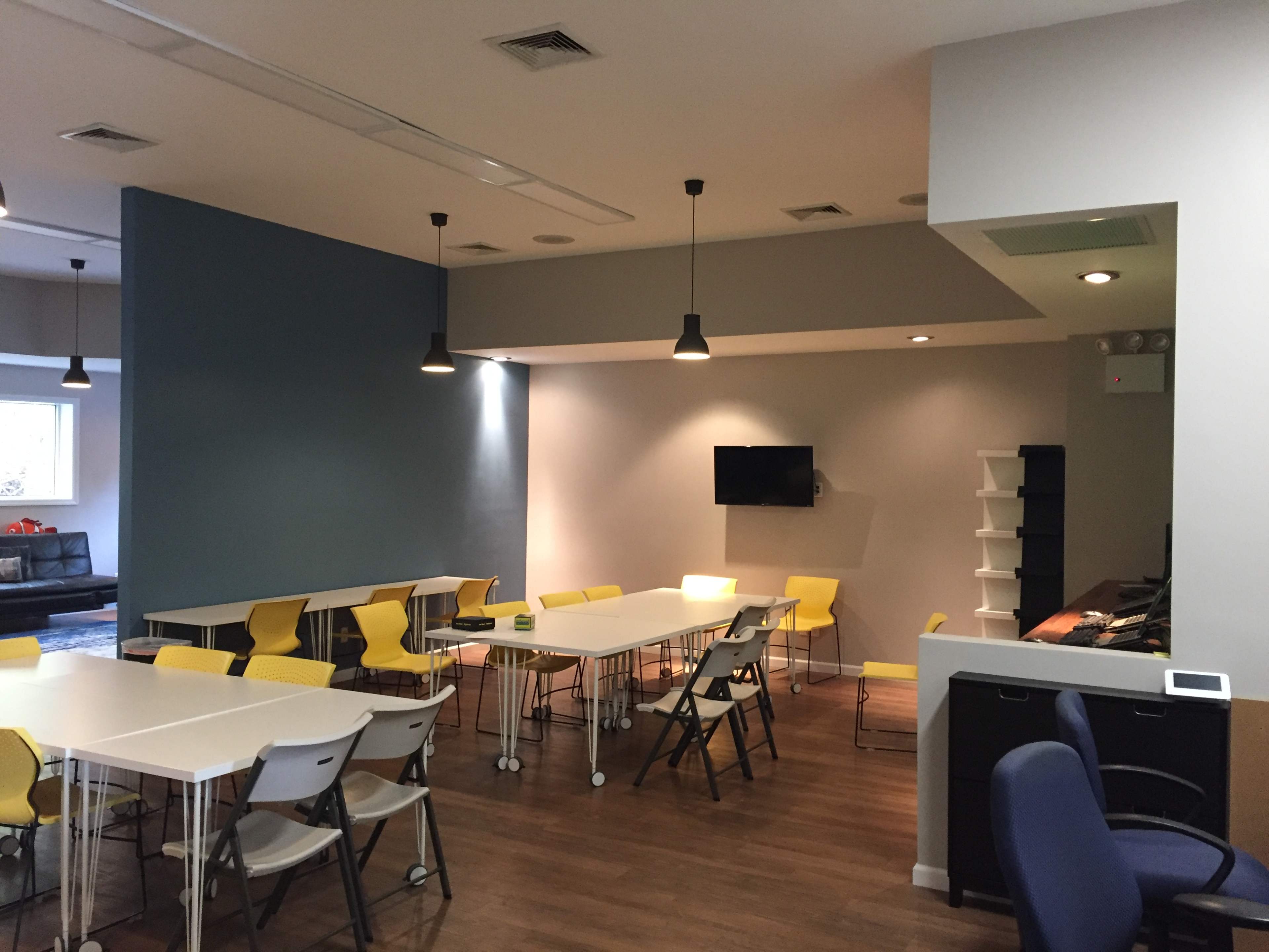 A modern meeting room with several tables and chairs, a large TV on the wall, and pendant lighting.