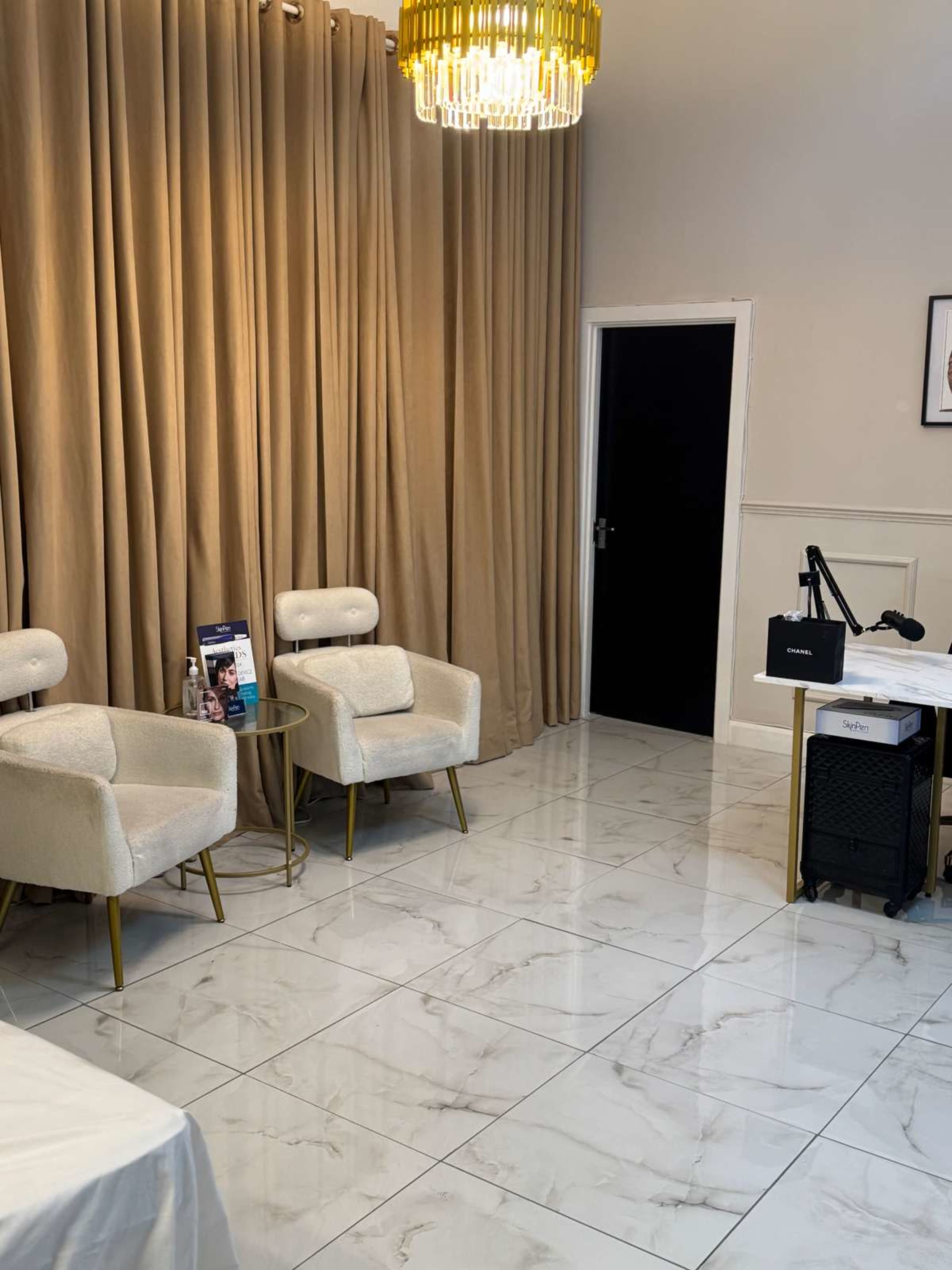 The image shows a modern room with beige curtains, two white upholstered chairs, a chandelier, and a desk with equipment in one corner.