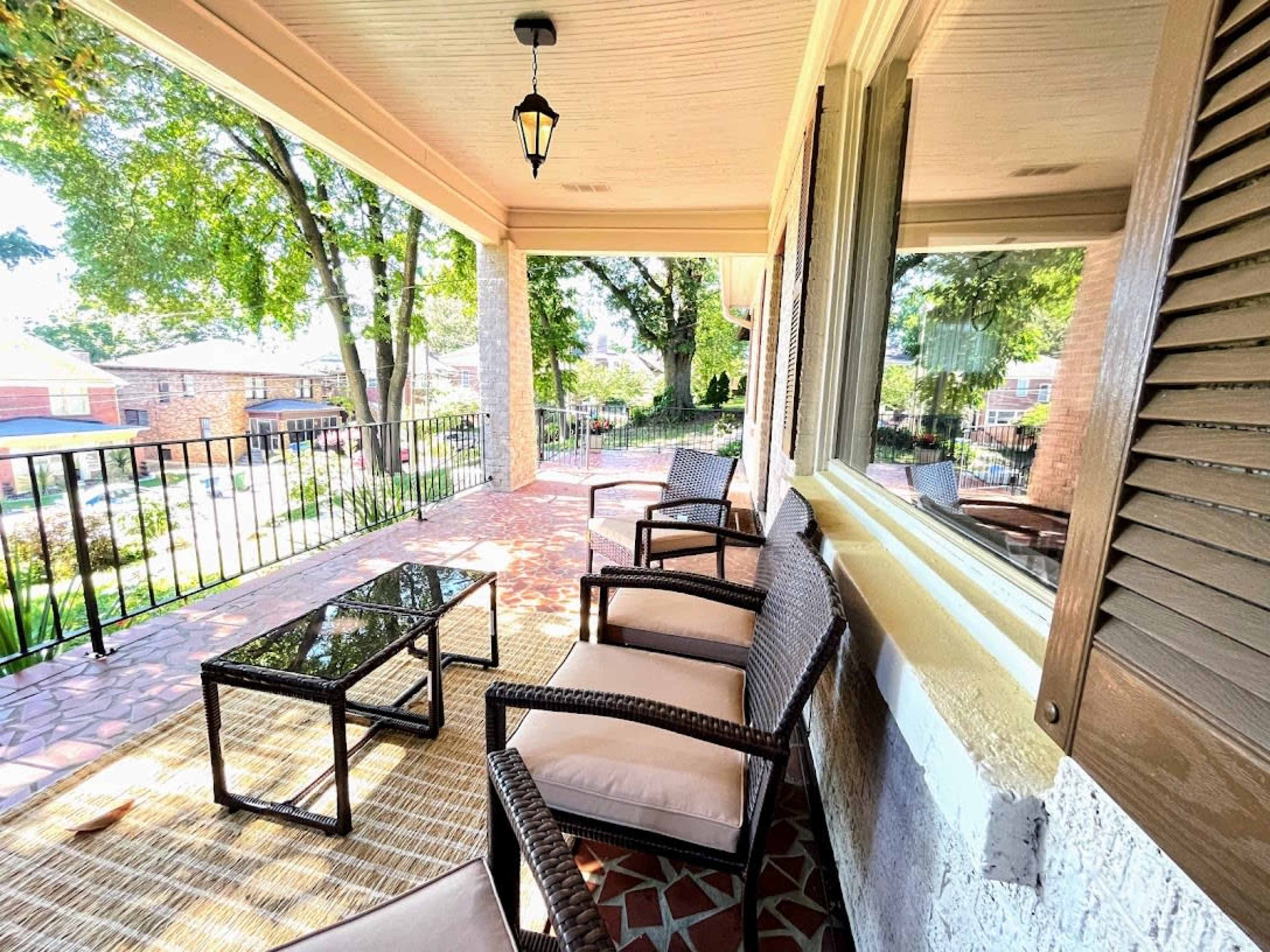 A covered porch with several chairs and a glass-top coffee table, surrounded by greenery and visible houses beyond a railing.