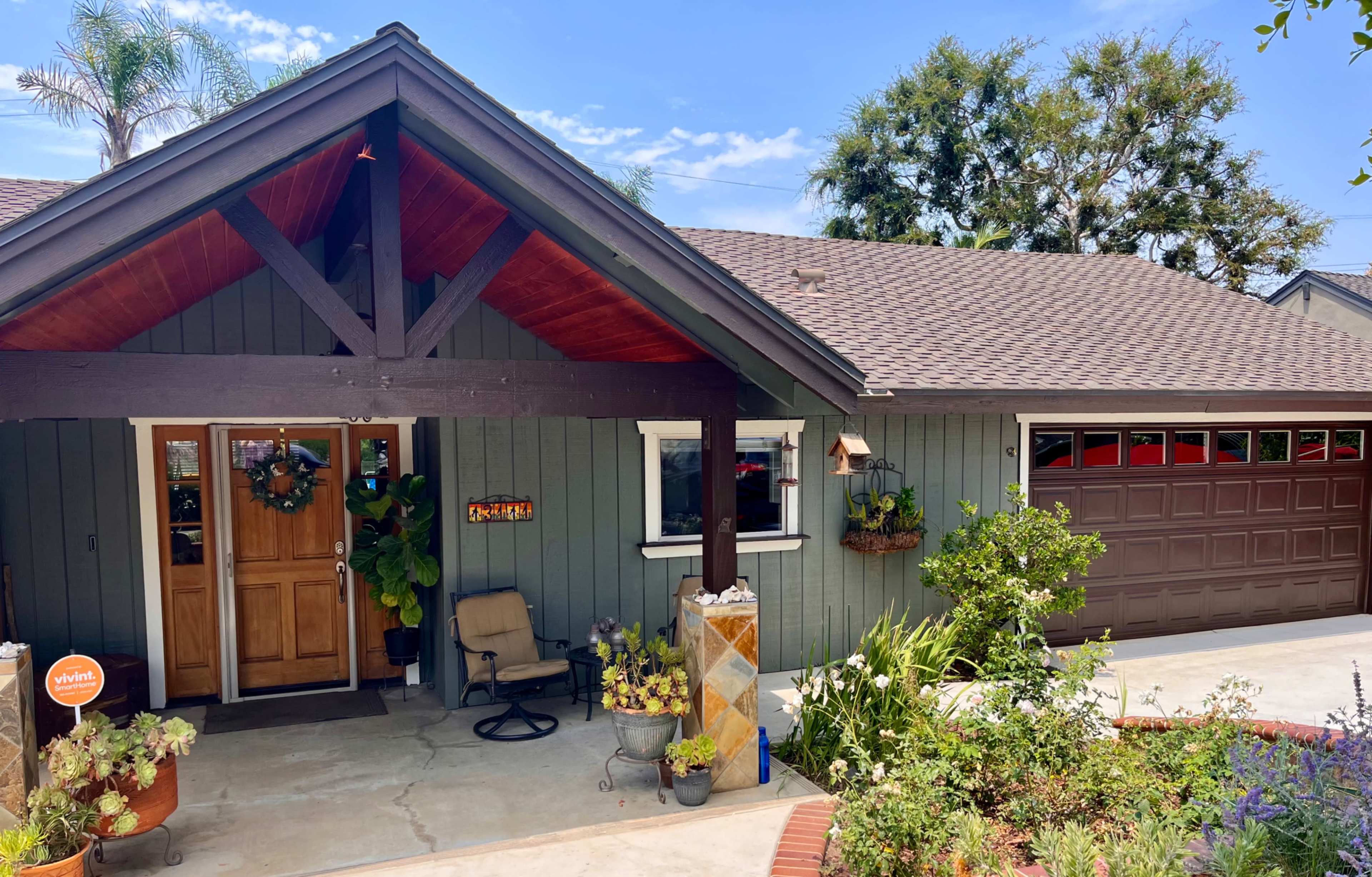 A single-story house with a wooden front porch, a double door entrance, and a garage on the right, surrounded by landscaped plants and a decorative planter.