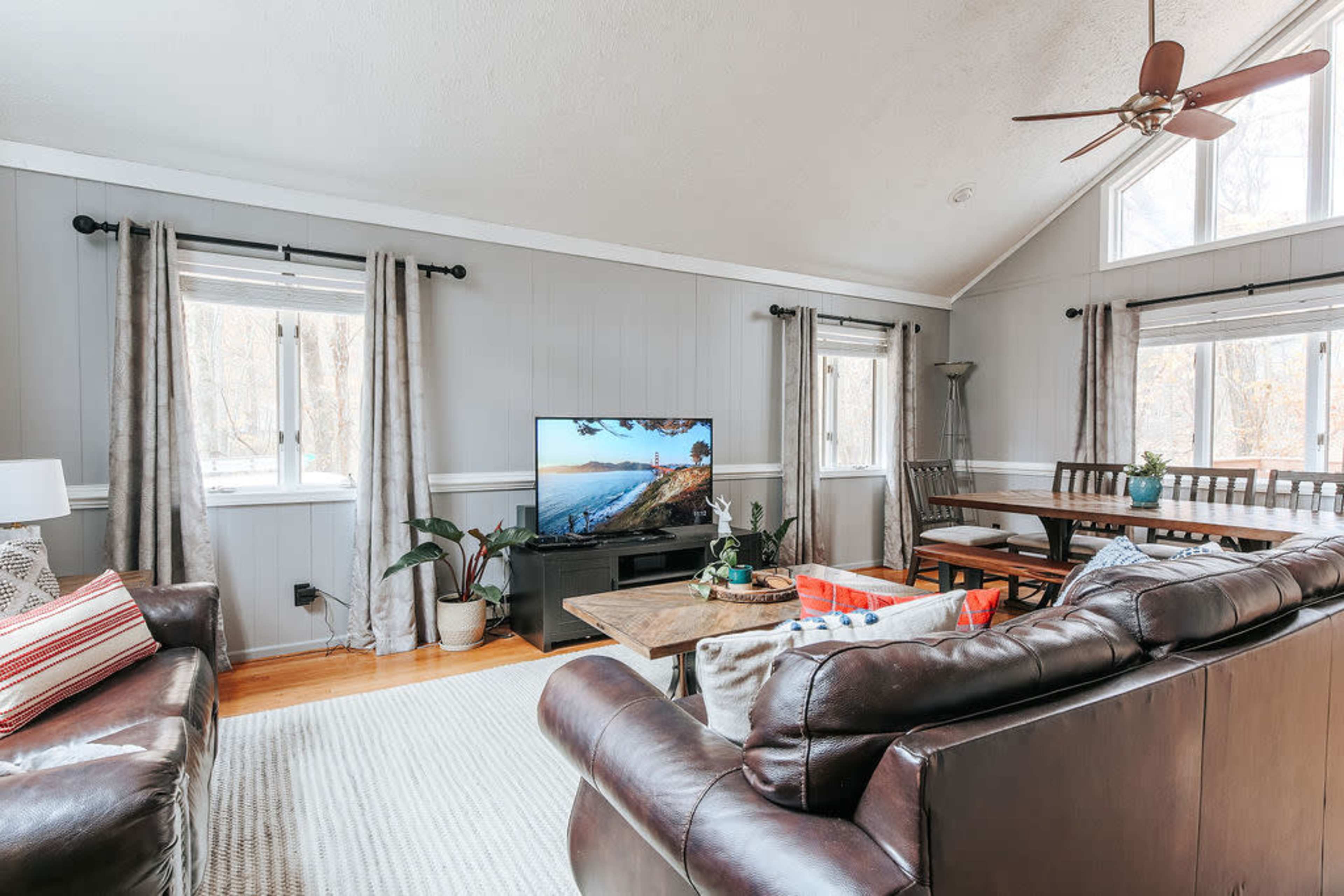 A cozy living room features a television mounted on a stand, two leather sofas, a dining area, and large windows allowing natural light in.