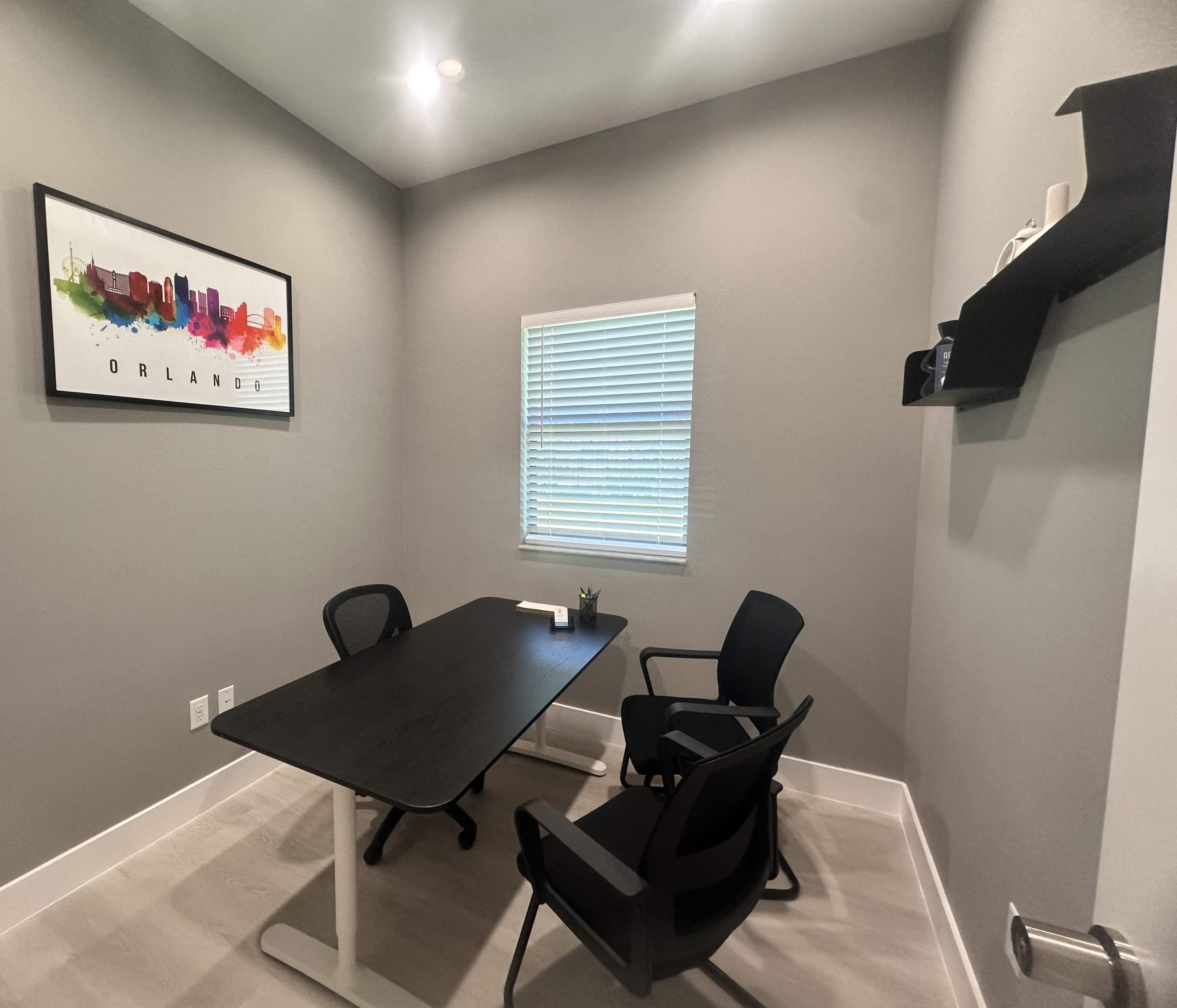 A small office with a black table and two chairs, featuring a window with white blinds and a colorful cityscape painting on the wall.