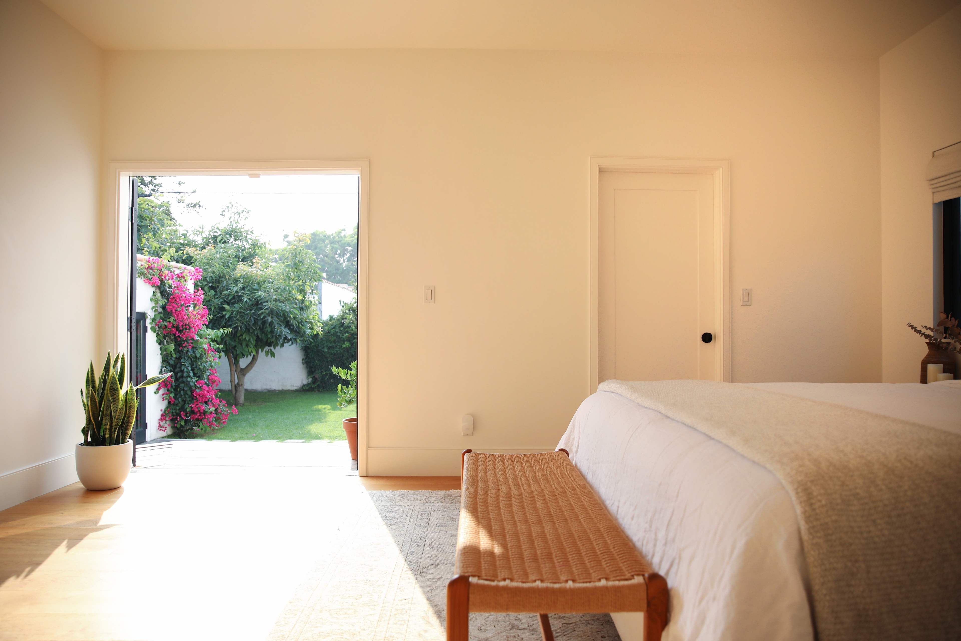 A bright bedroom features a bed with a light-colored coverlet, a wooden bench at its foot, and a door that opens to a garden with blooming plants.