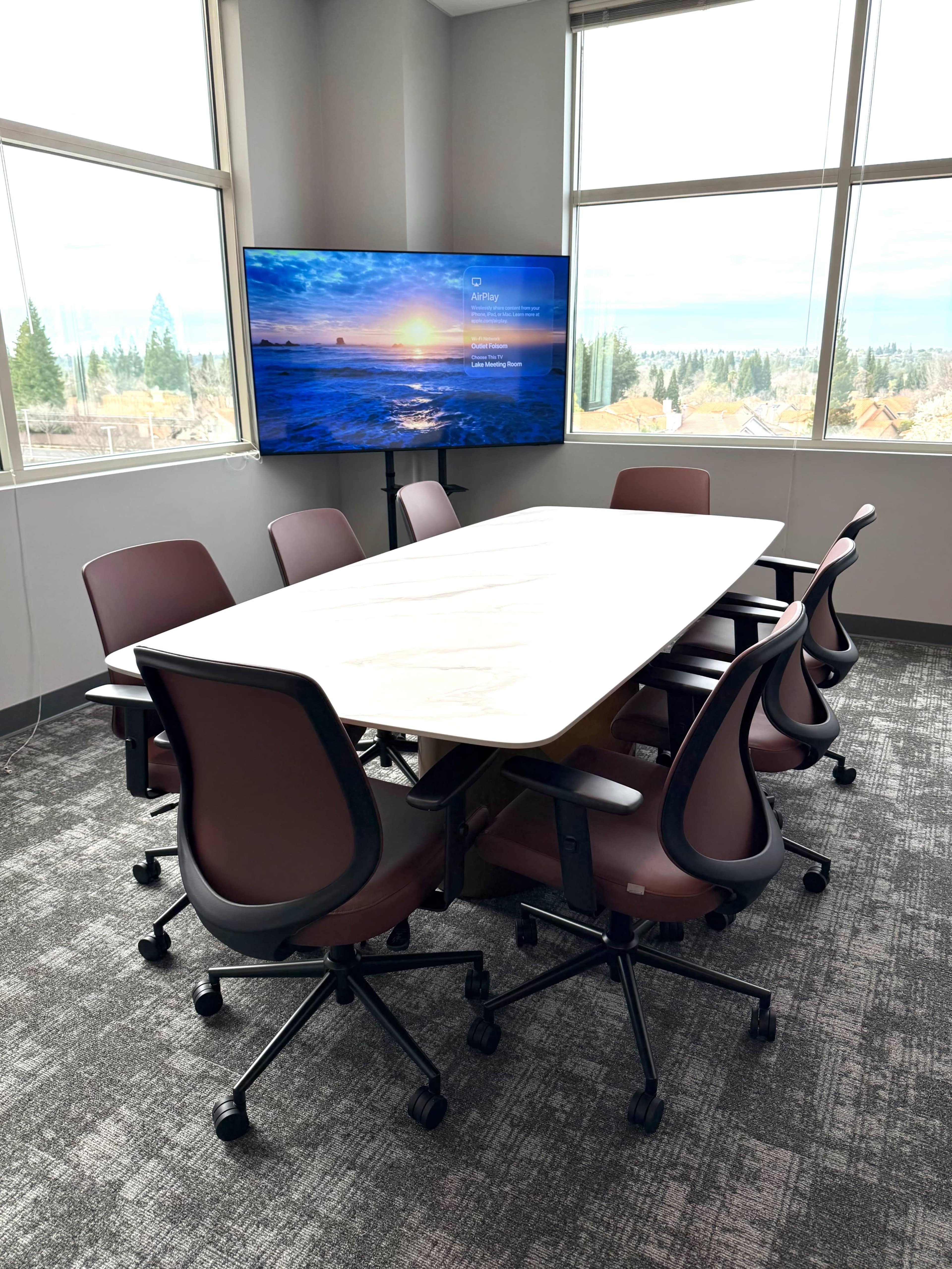 A conference room features a large table surrounded by six brown chairs and a television displaying a scenic sunset.