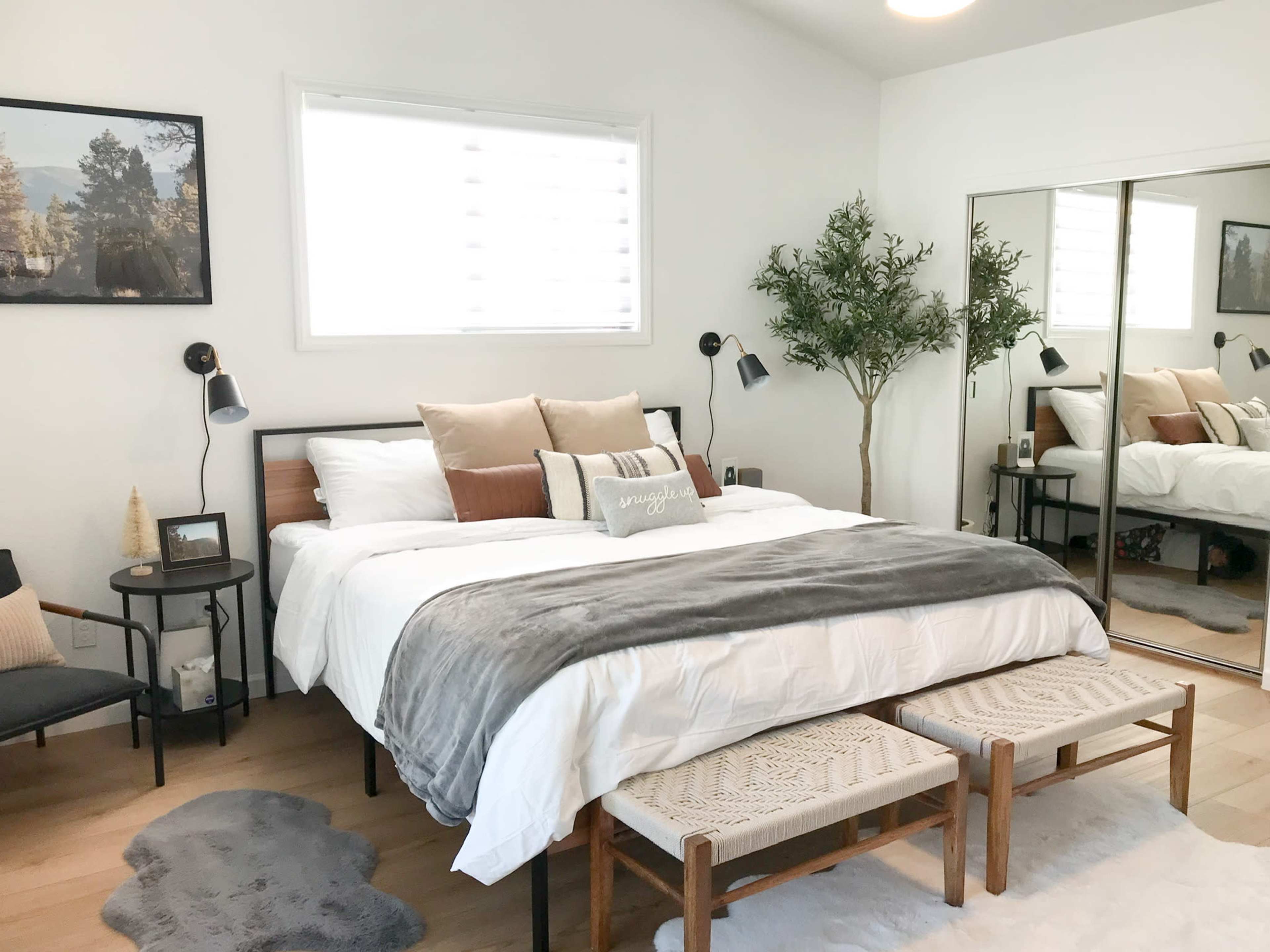 The image shows a modern bedroom featuring a large bed with white linens, two decorative pillows, and a pair of woven benches at the foot, along with a mirror and a small plant.