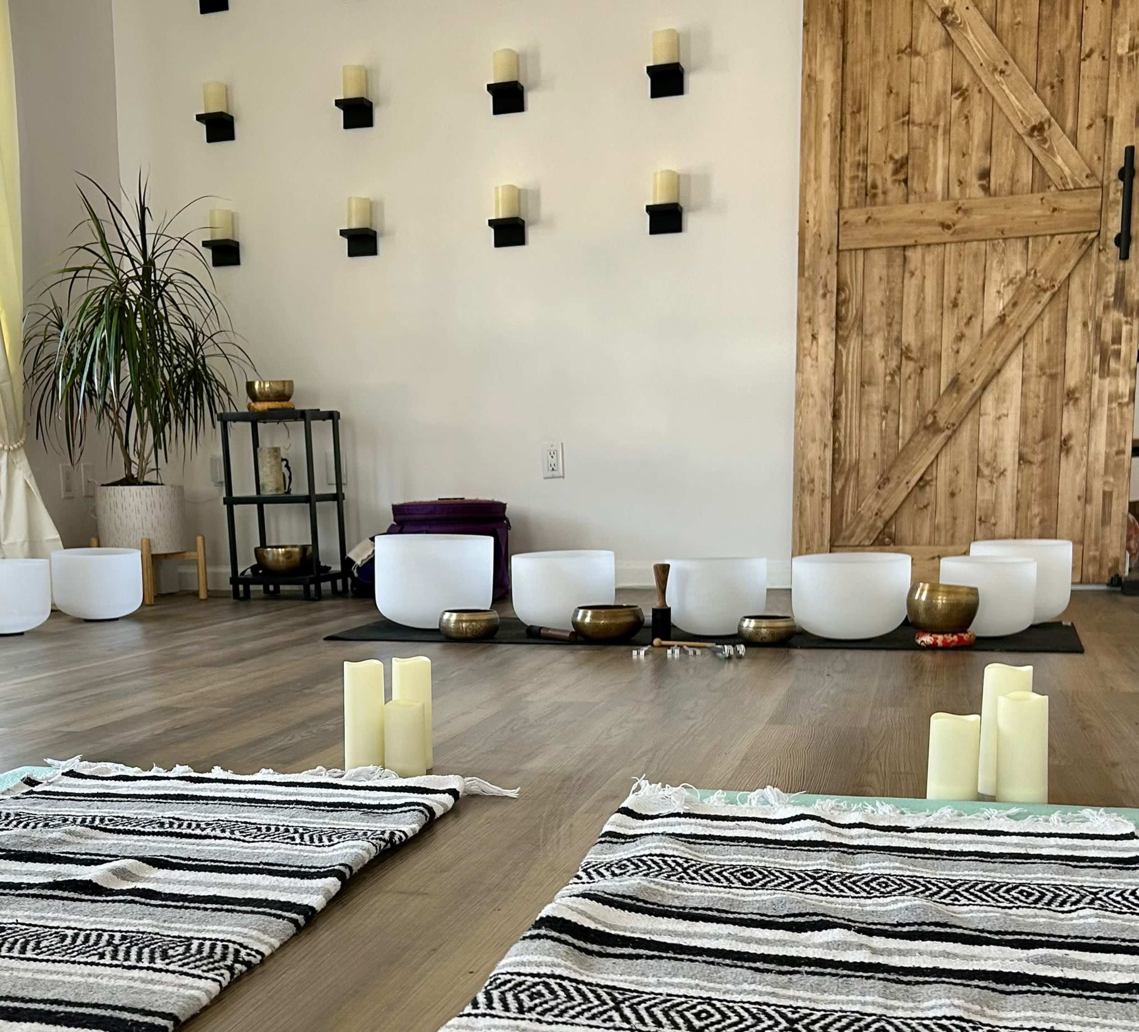 A spacious room with two patterned rugs, several crystal singing bowls arranged on one side, a wooden door, and wall-mounted candle holders.