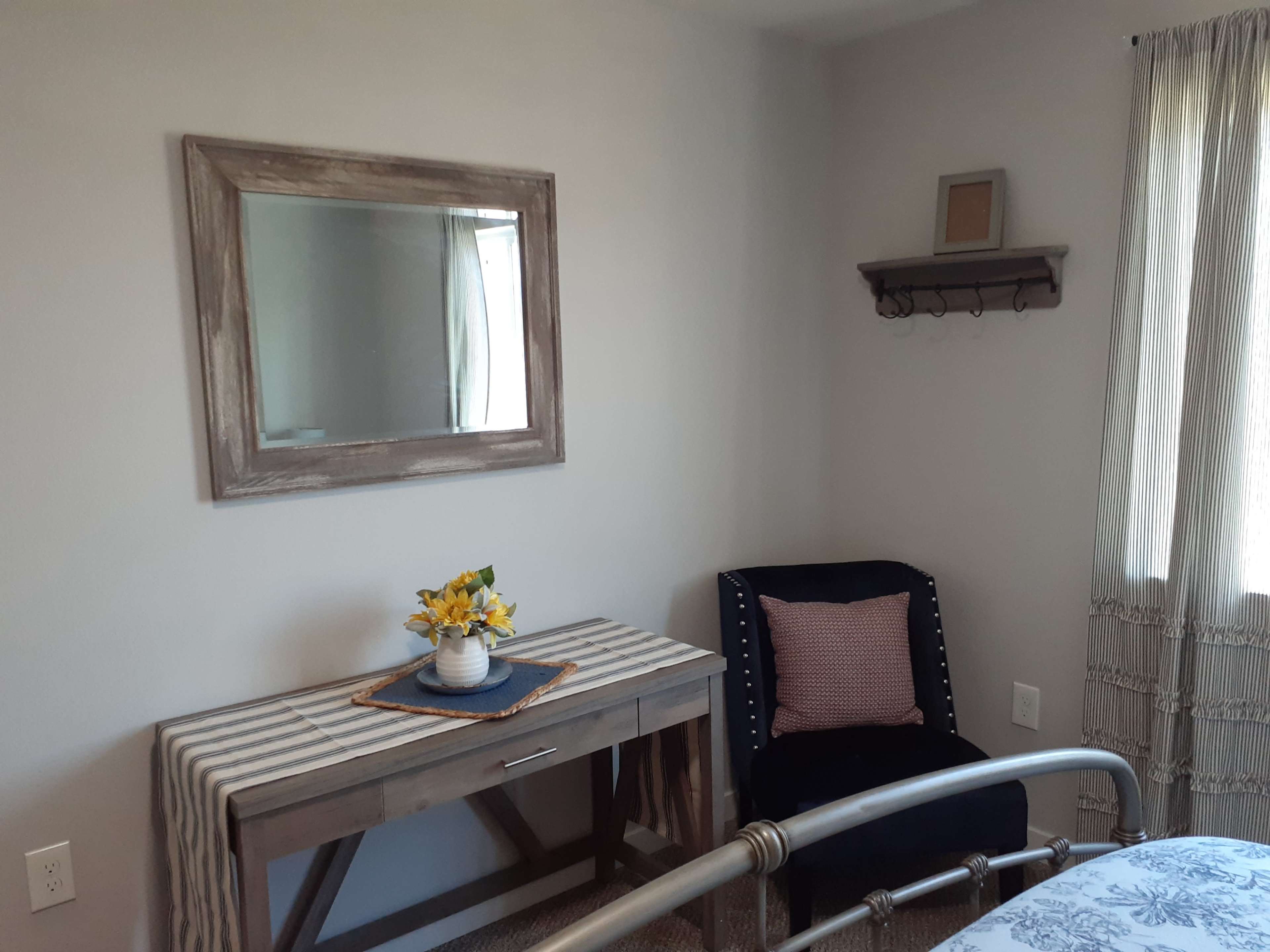 A small bedroom with a table and mirror on one wall, a navy blue chair in the corner, and a window with sheer curtains.