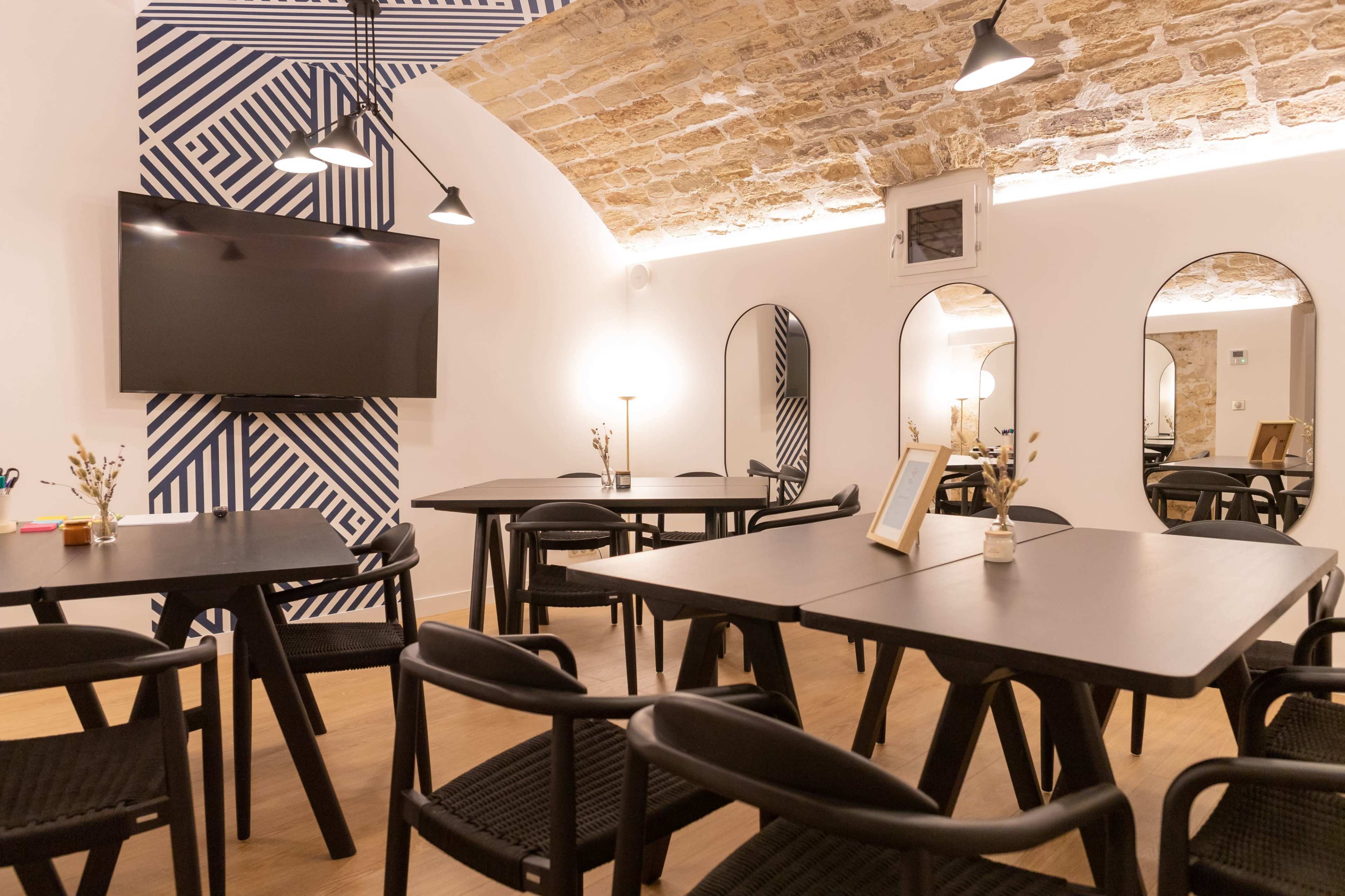 A modern meeting room with dark wooden tables, black chairs, and a large wall-mounted TV, complemented by stone archways and decorative mirrors.