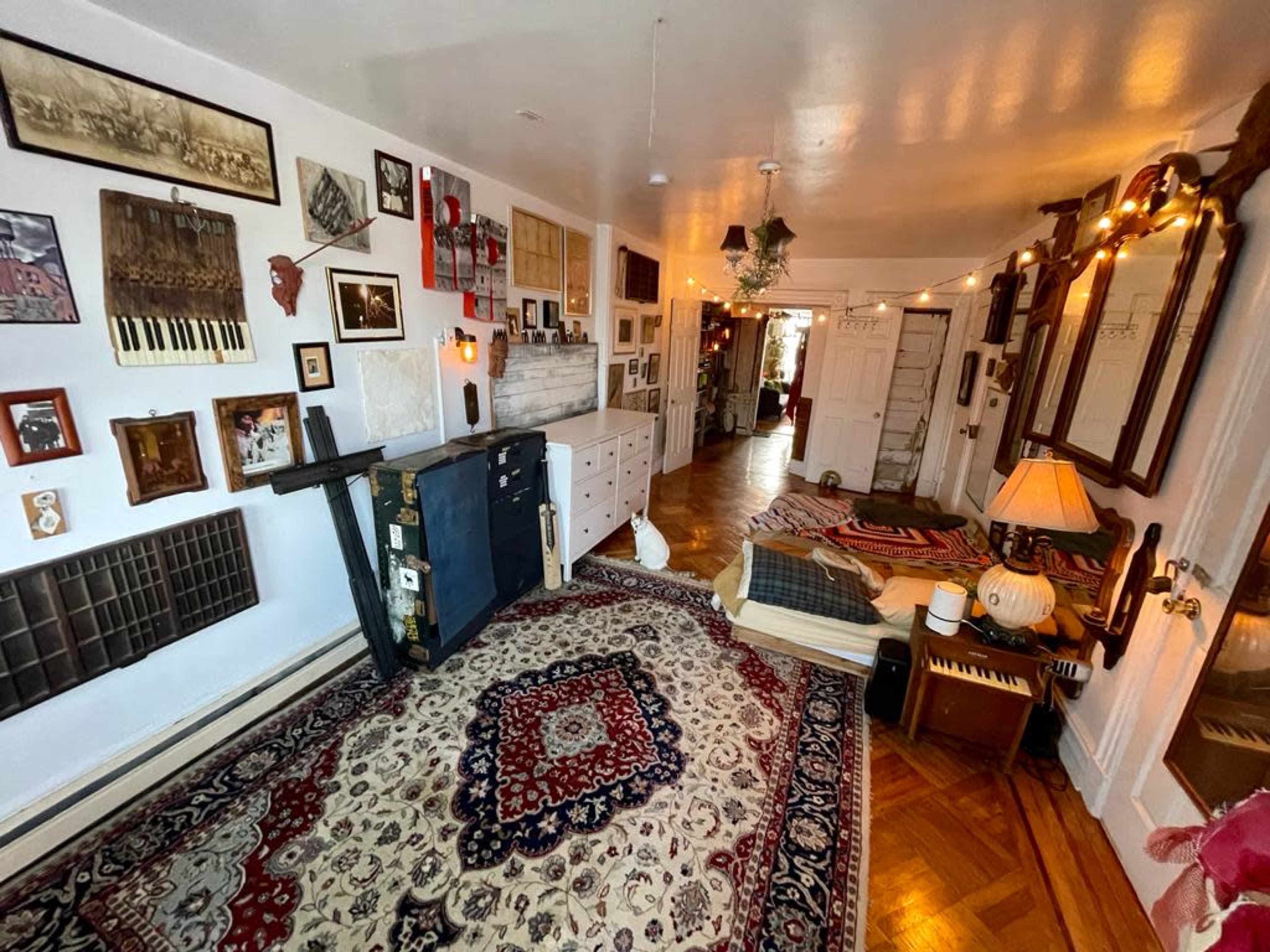 A cozy room features a patterned rug, a mix of framed artwork on the walls, a wooden bed with layered bedding, and various decorative items scattered throughout.