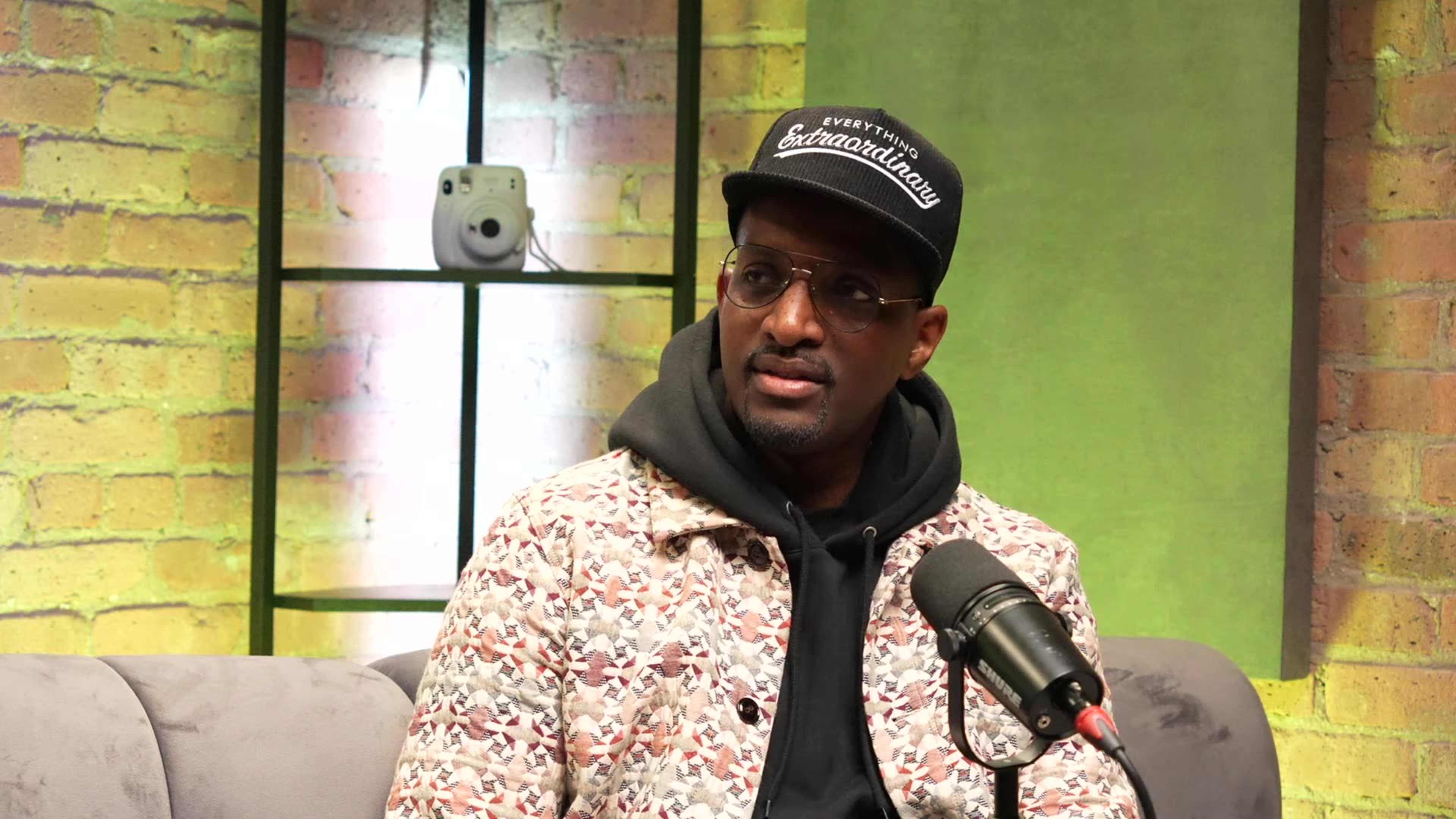 A man in a patterned jacket and black cap sits in front of a microphone against a backdrop of illuminated shelves.