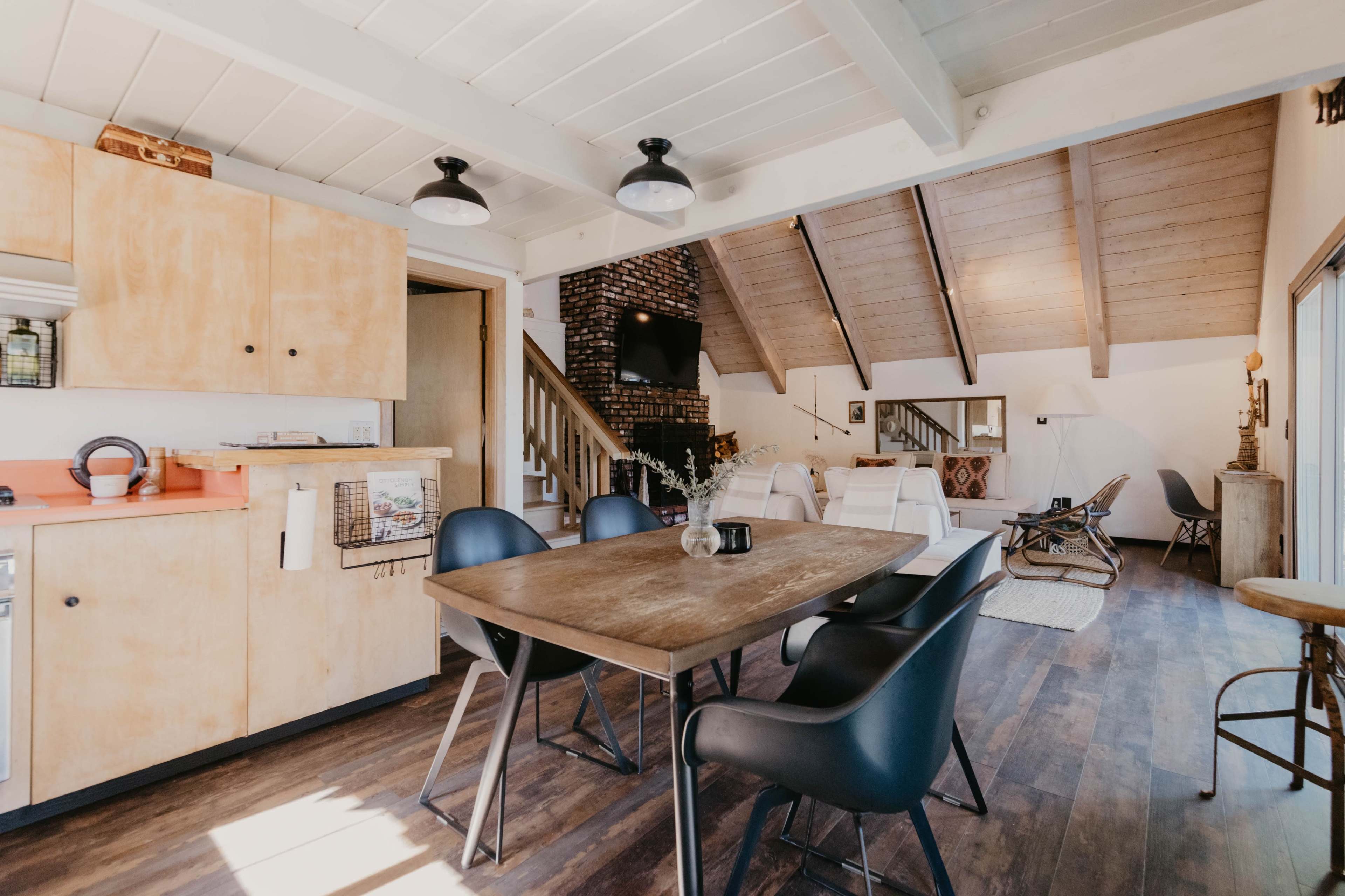 A cozy, modern kitchen and dining area features a wooden table surrounded by black chairs, with light-colored cabinetry and exposed beams overhead.