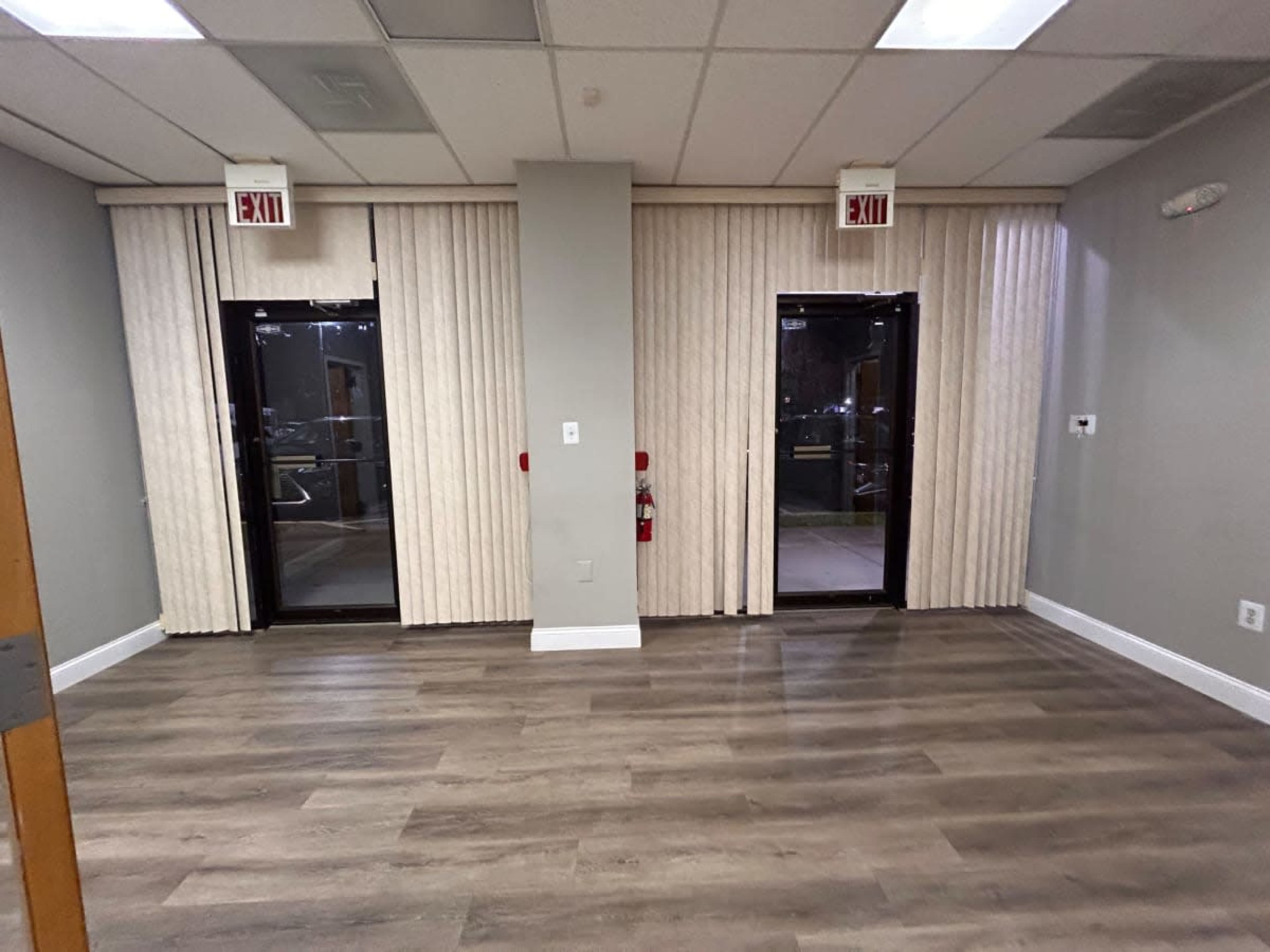 A sparsely furnished room with light-colored walls, two exit doors on opposite sides, and a wooden floor.