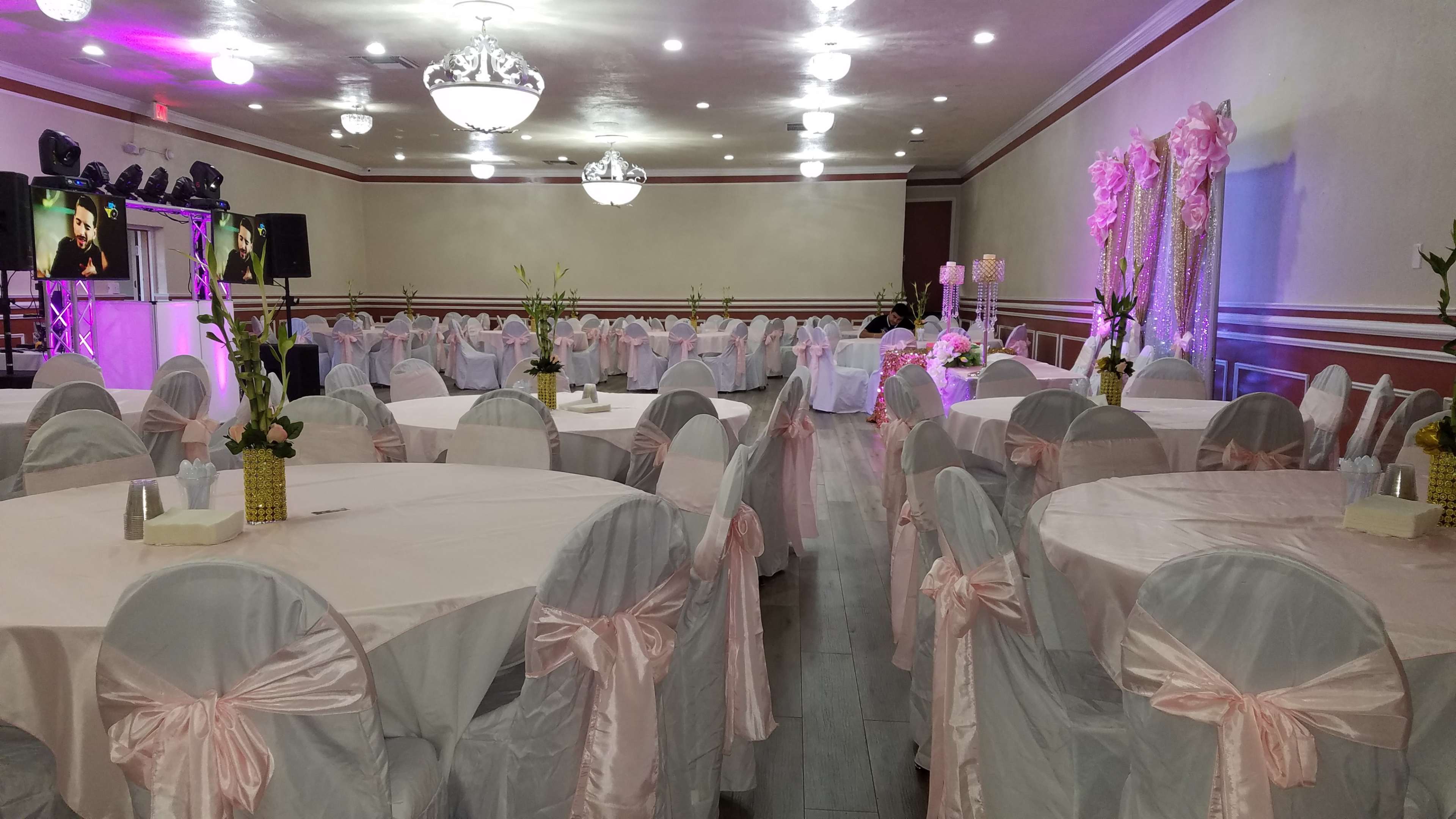 The image shows a large banquet hall set up for an event, featuring round tables with pink tablecloths and white chairs adorned with pink sashes.