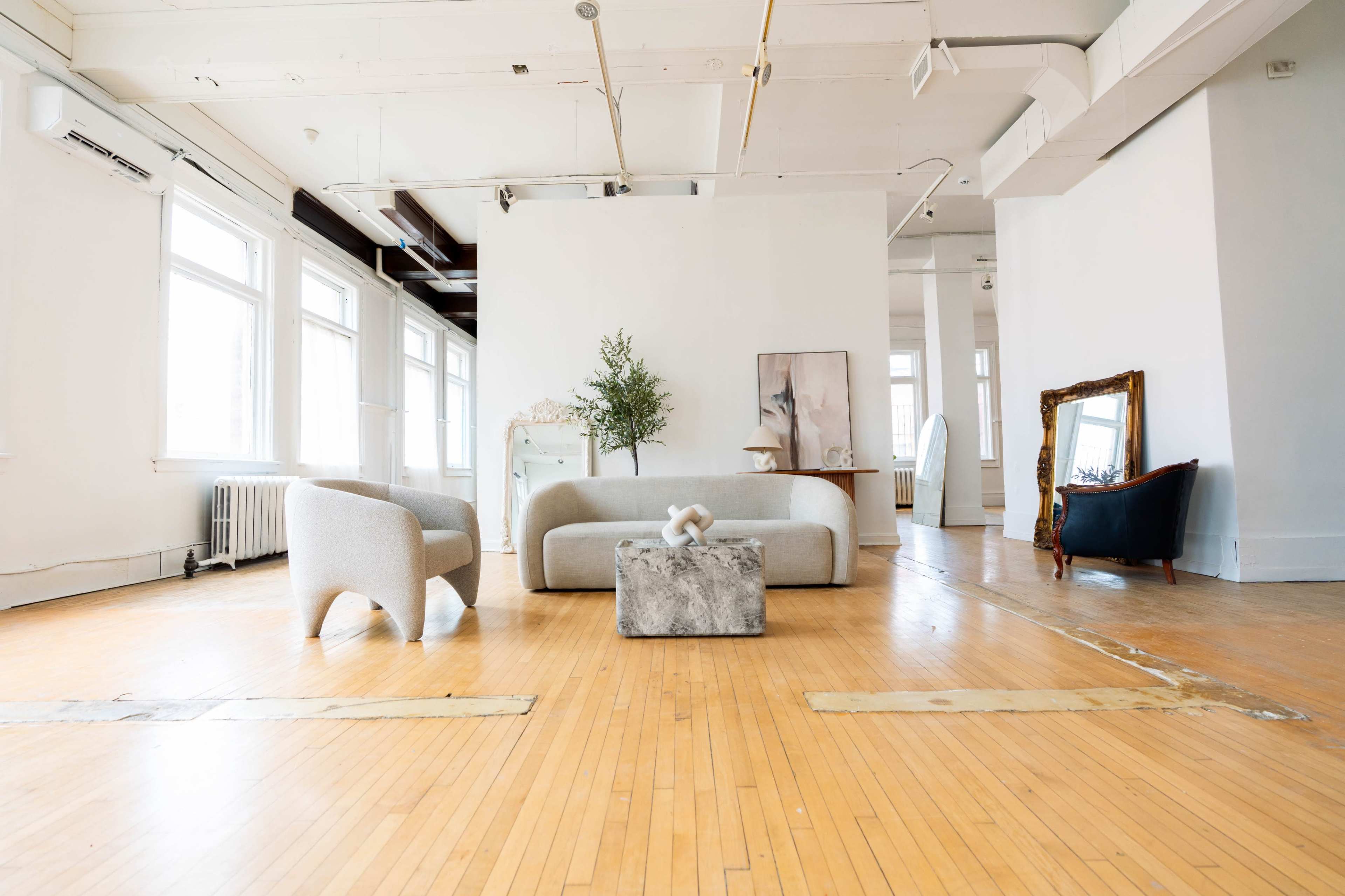 The image shows a spacious, light-filled room with a minimalist sofa and a coffee table placed on a wooden floor, along with a few decorative elements and furniture pieces.