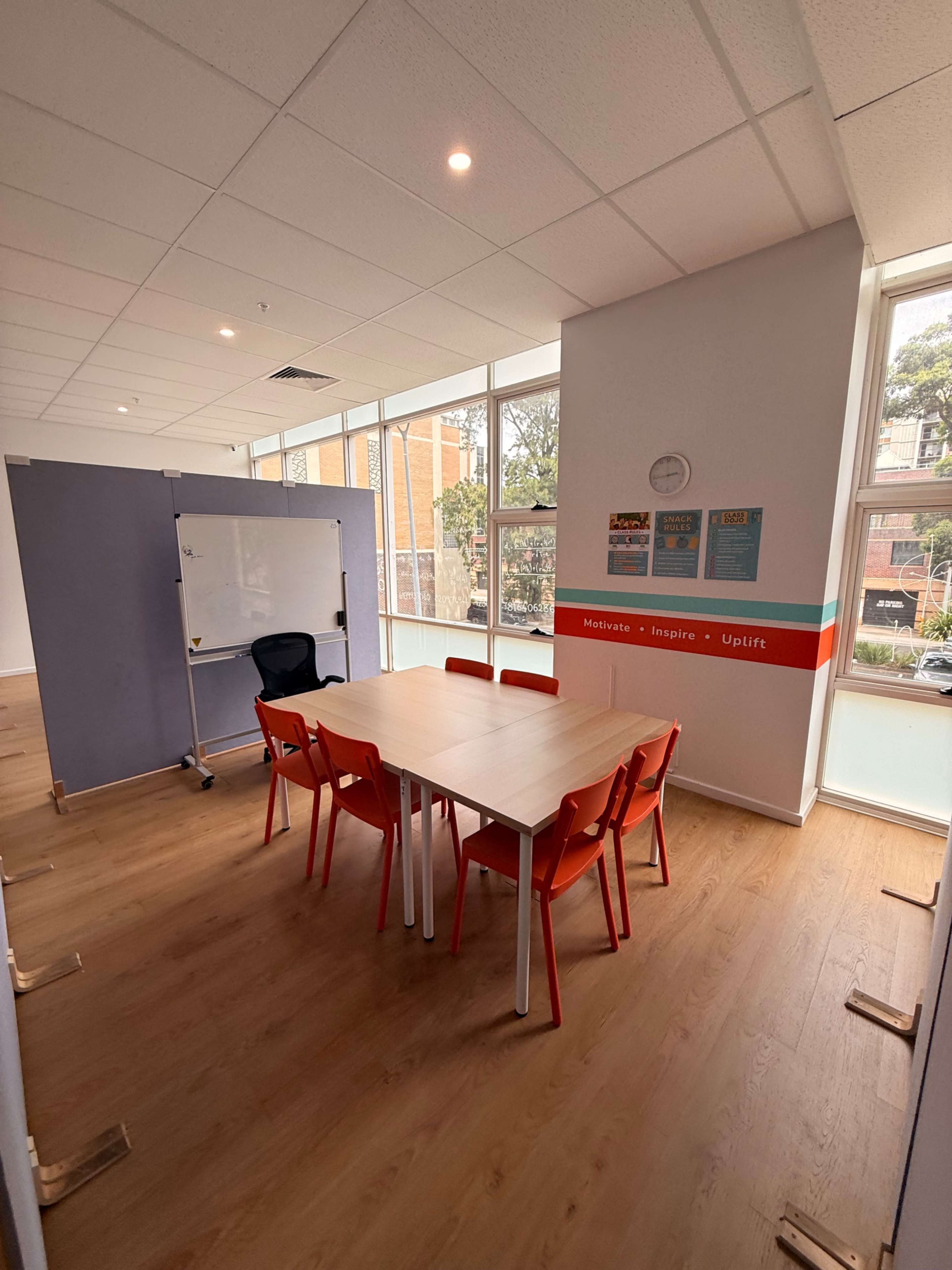 The image shows a modern meeting room with a wooden floor, a large table surrounded by red chairs, a whiteboard on a movable partition, and large windows letting in natural light.