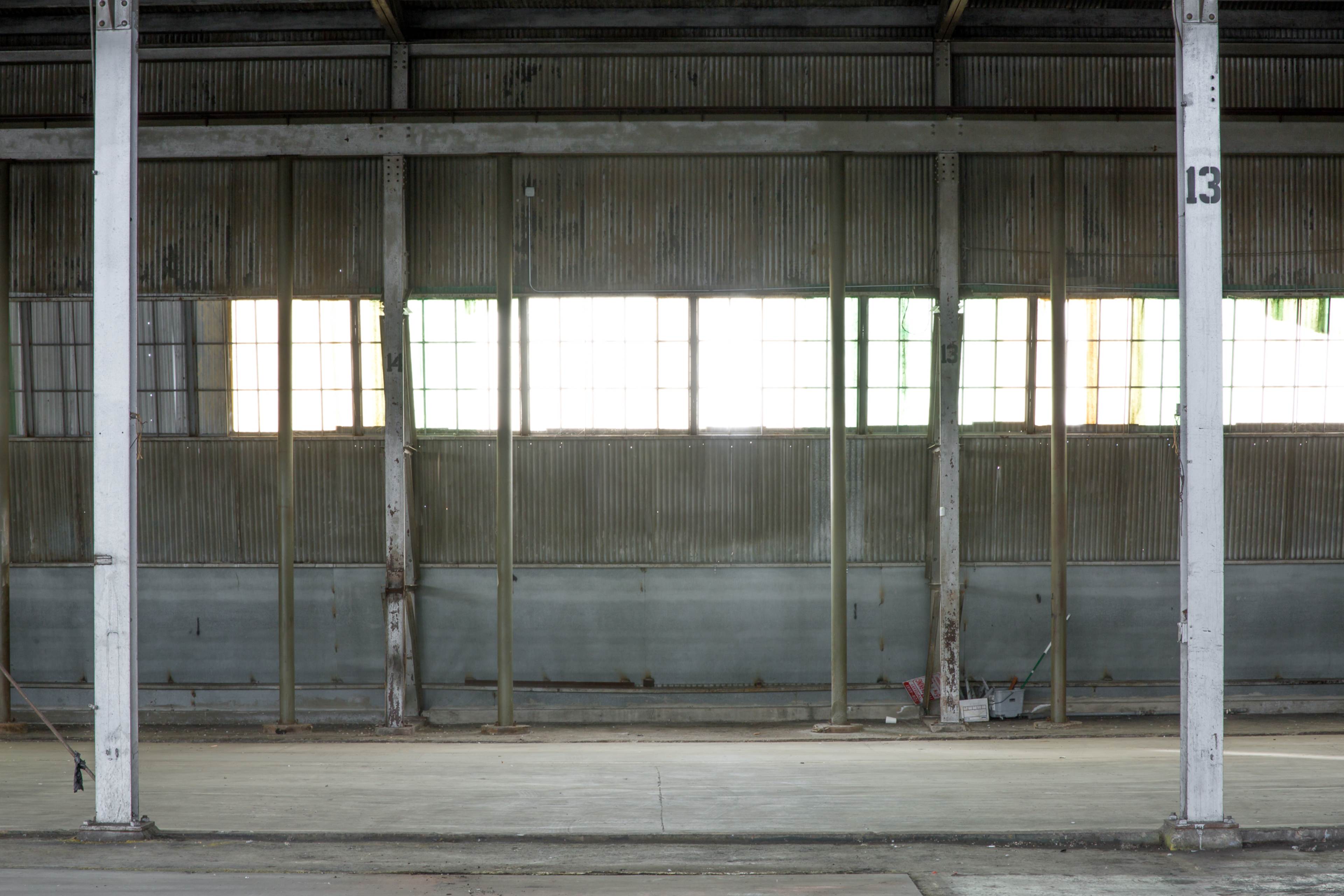 The interior of a large, empty industrial building with metal walls and windows, featuring visible support columns and a concrete floor.