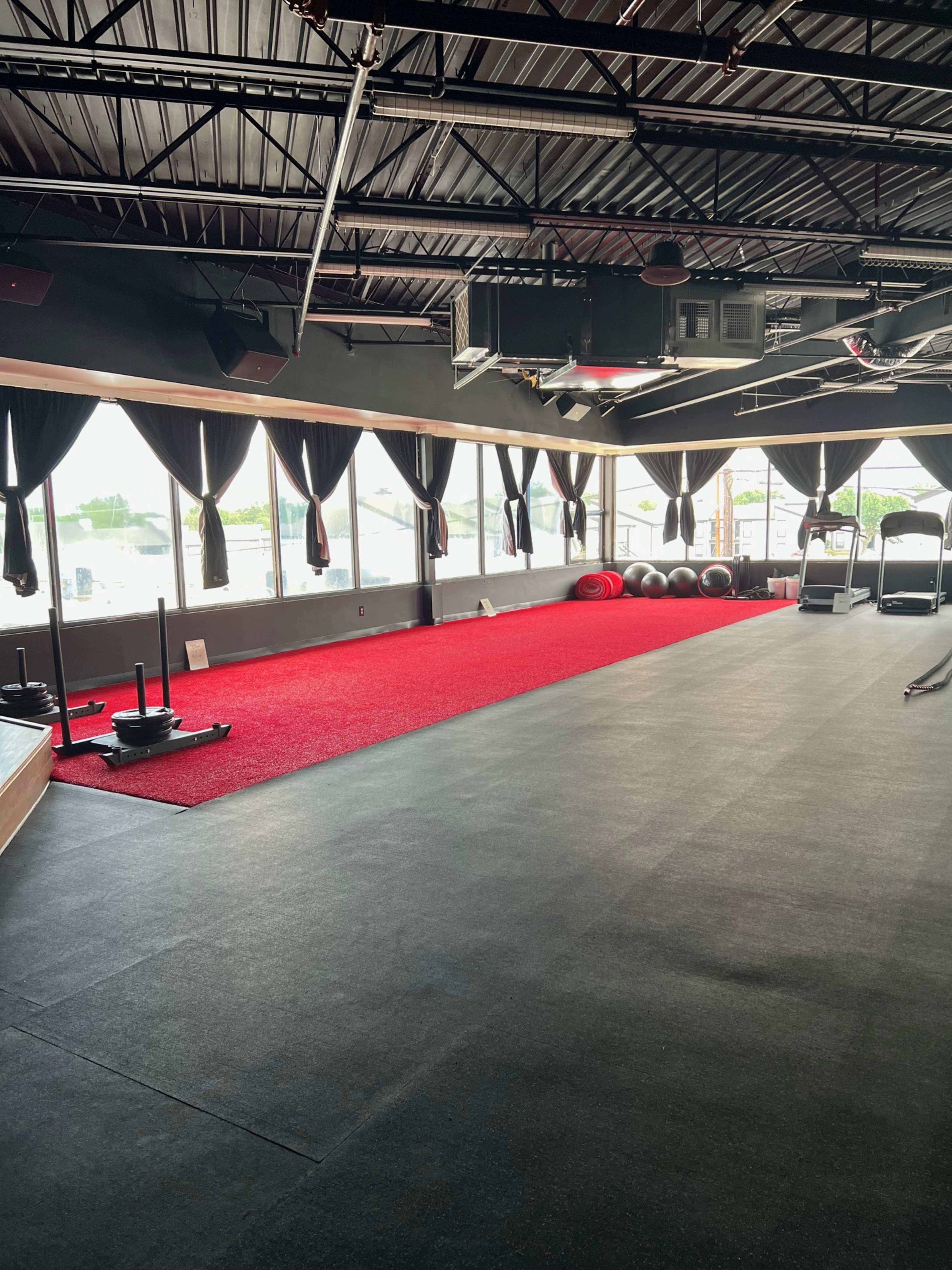 A fitness studio with red carpet flooring, workout equipment, and large windows covered by black curtains.