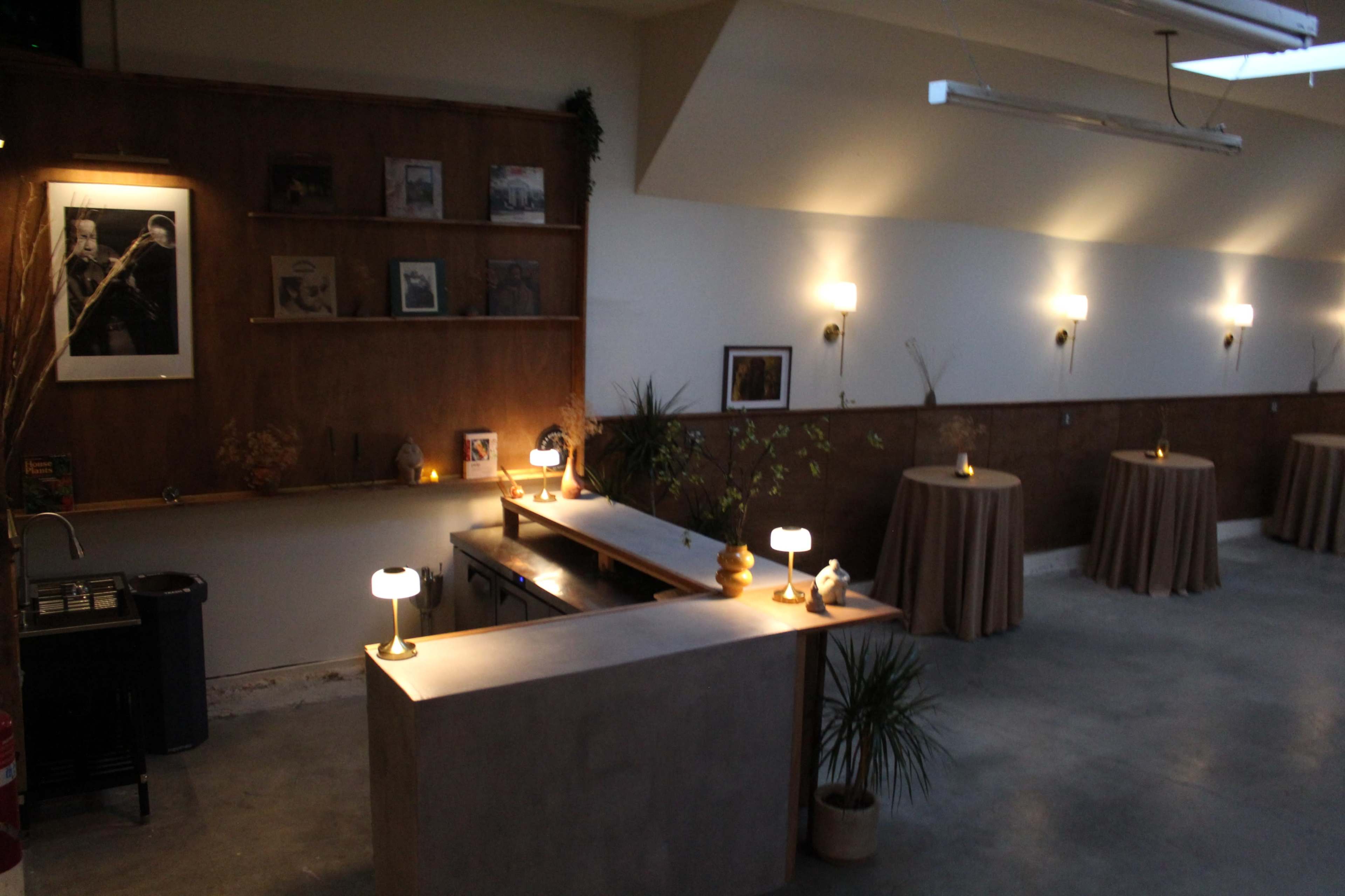 The image shows a modern interior space with a concrete bar, tapered tables covered in beige cloth, and walls adorned with framed pictures and warm lighting.