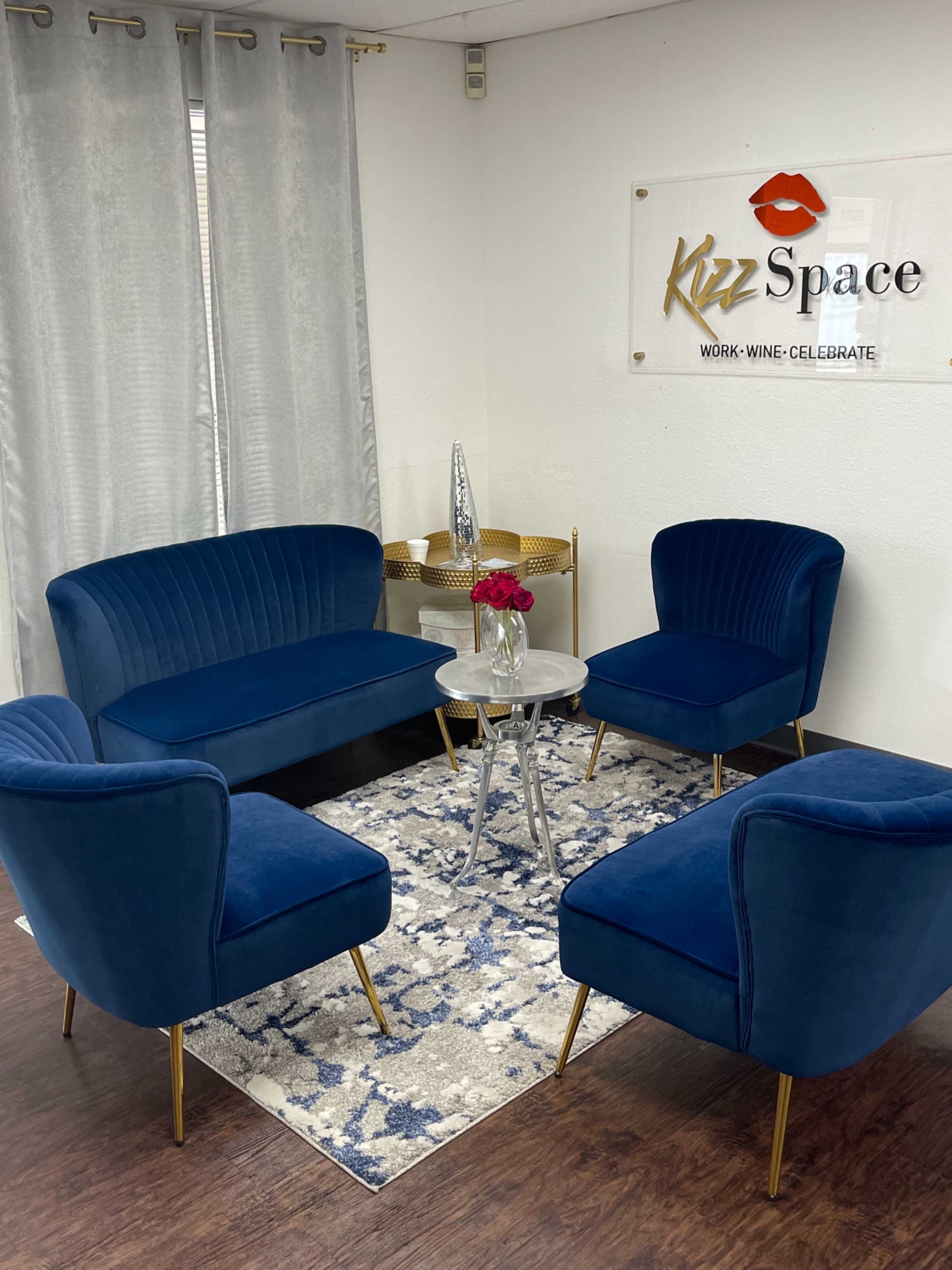 The image shows a small, modern seating area featuring a blue velvet sofa and chairs arranged around a glass coffee table, placed on a patterned rug against a white wall with a decorative sign.