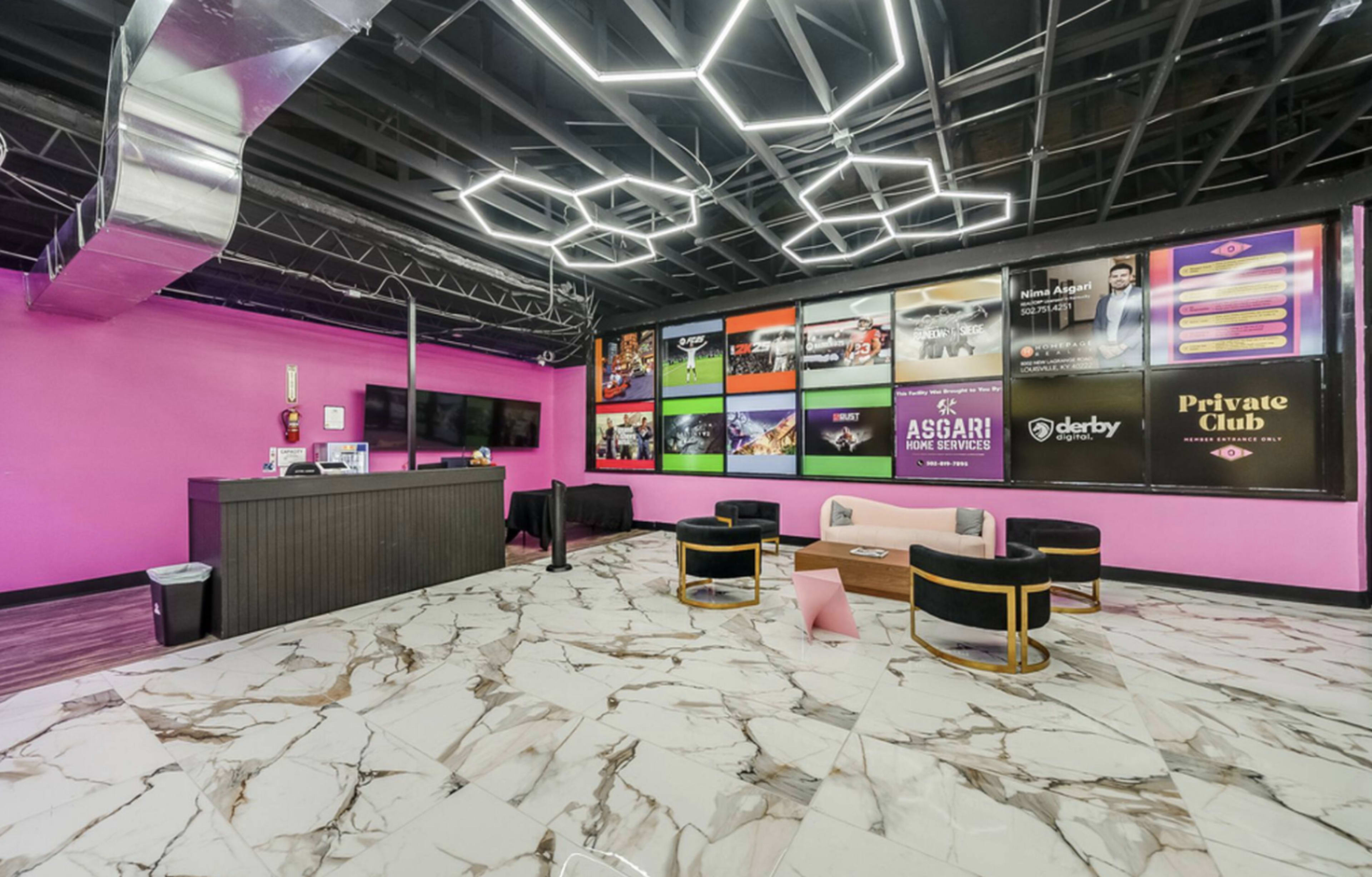 The image shows a modern lounge area with a pink accent wall, sleek furniture, and a display of promotional screens mounted on the wall.