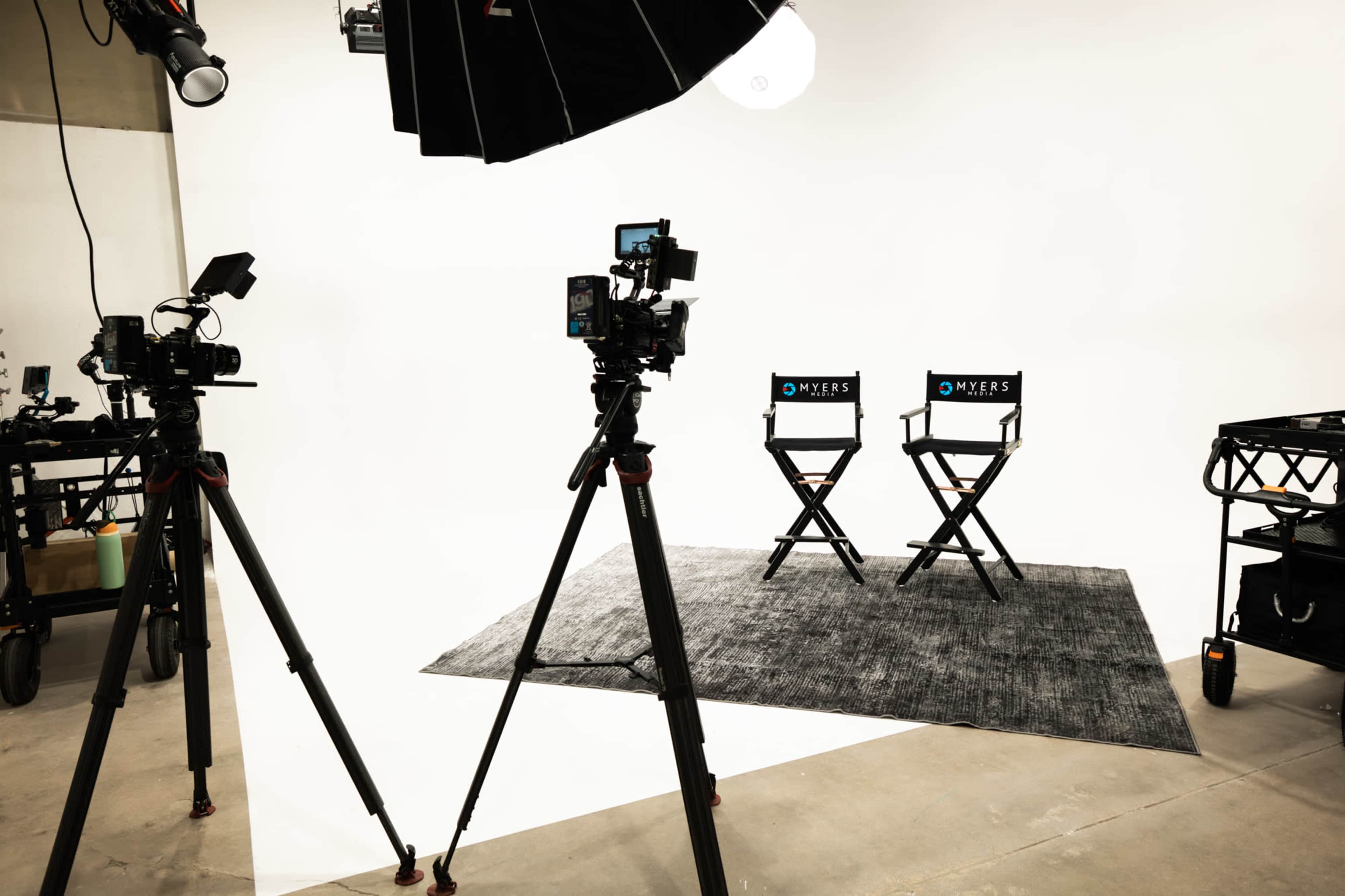 The image shows a studio setup with two director's chairs featuring the name "MYERS" and cameras mounted on tripods facing the chairs against a white backdrop.