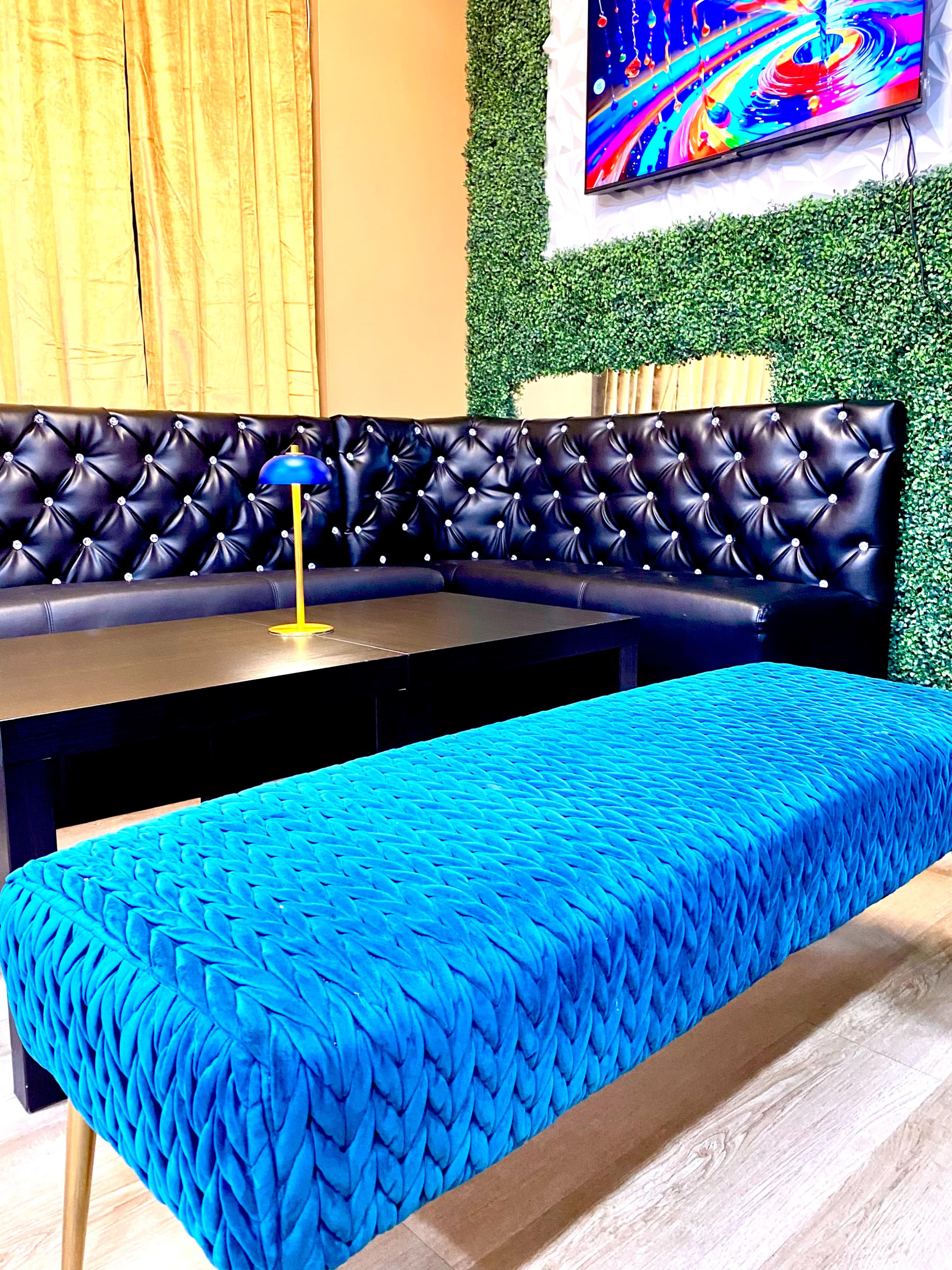 The image shows a modern seating area with a black tufted couch, a bright blue woven bench, and a television mounted on a wall covered with green foliage.