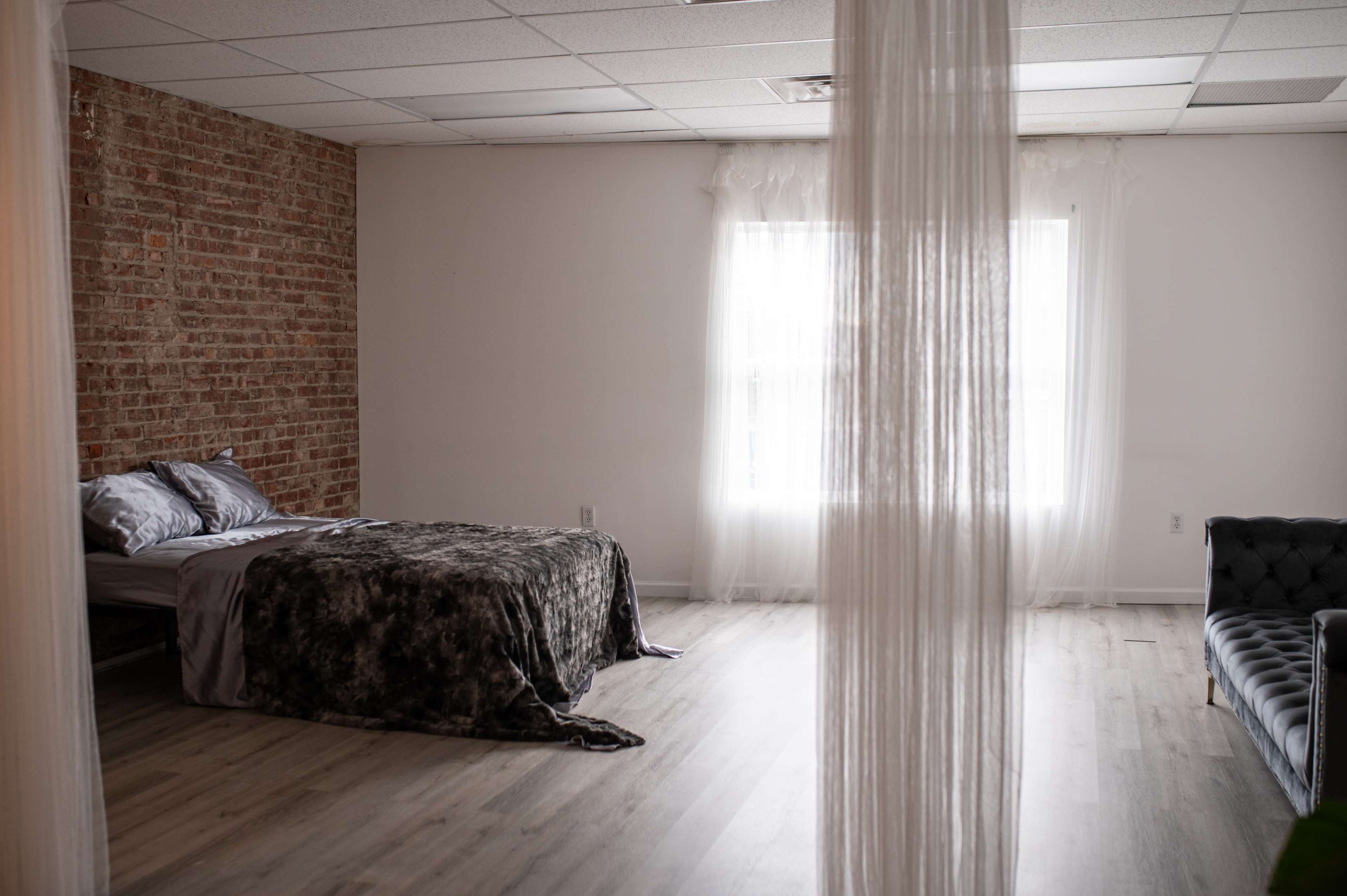 A minimalist bedroom with a bed covered in a dark blanket, a brick wall, and a large window with sheer curtains.