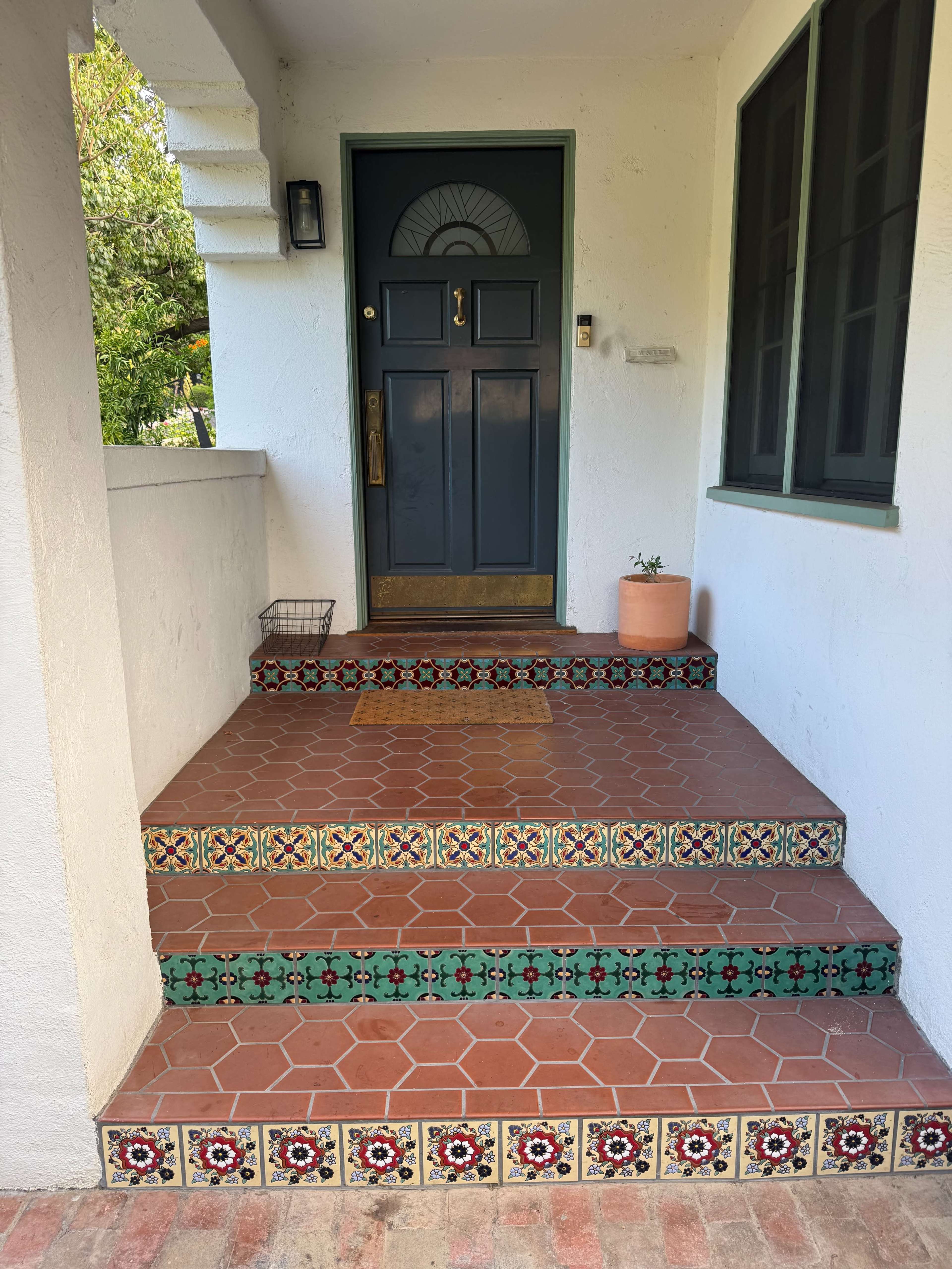 The image shows a front entrance with patterned tile steps, a dark blue door, and a small potted plant next to the door.