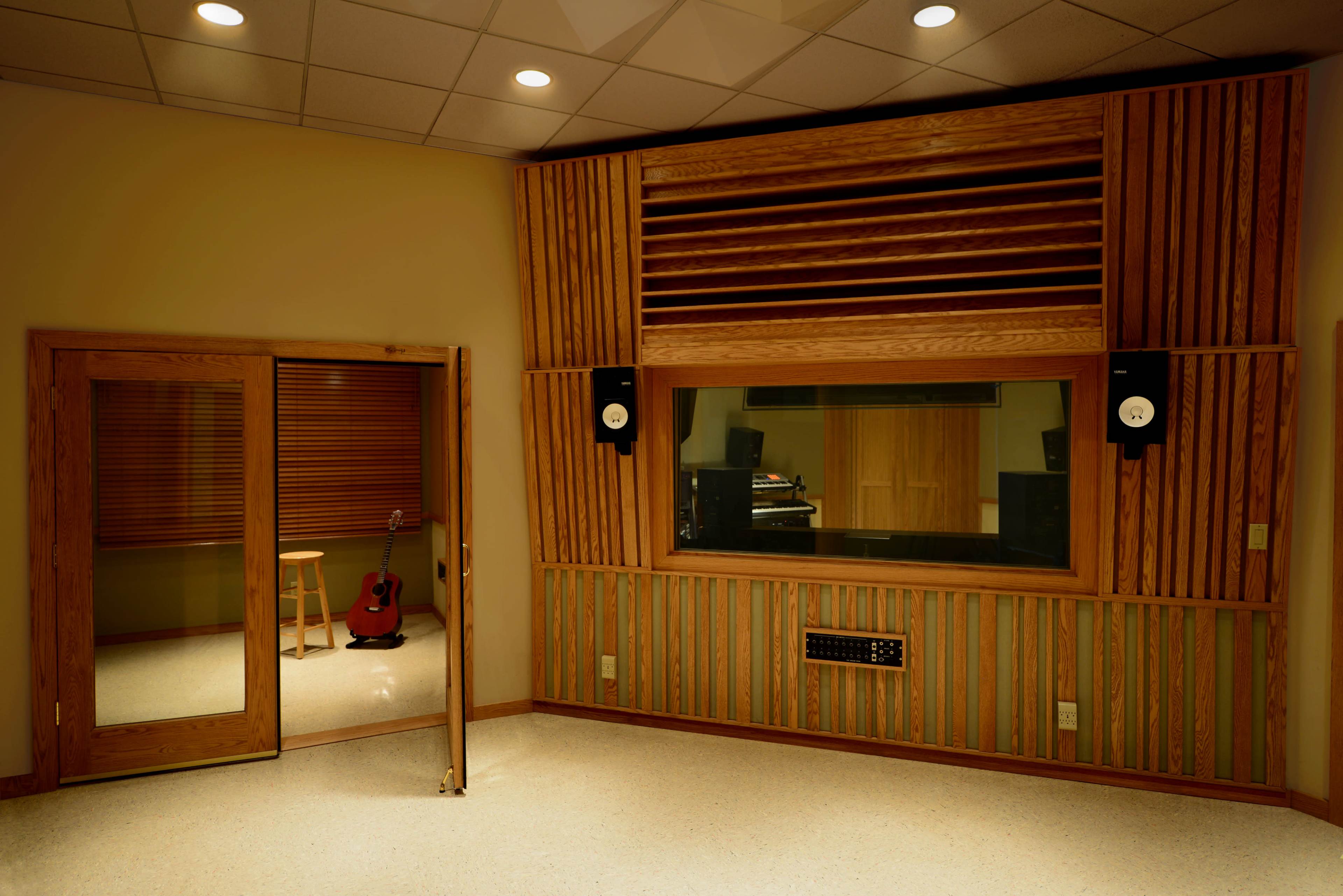 A sound recording studio featuring wooden acoustic panels, a large glass window, and a guitar positioned next to a stool.