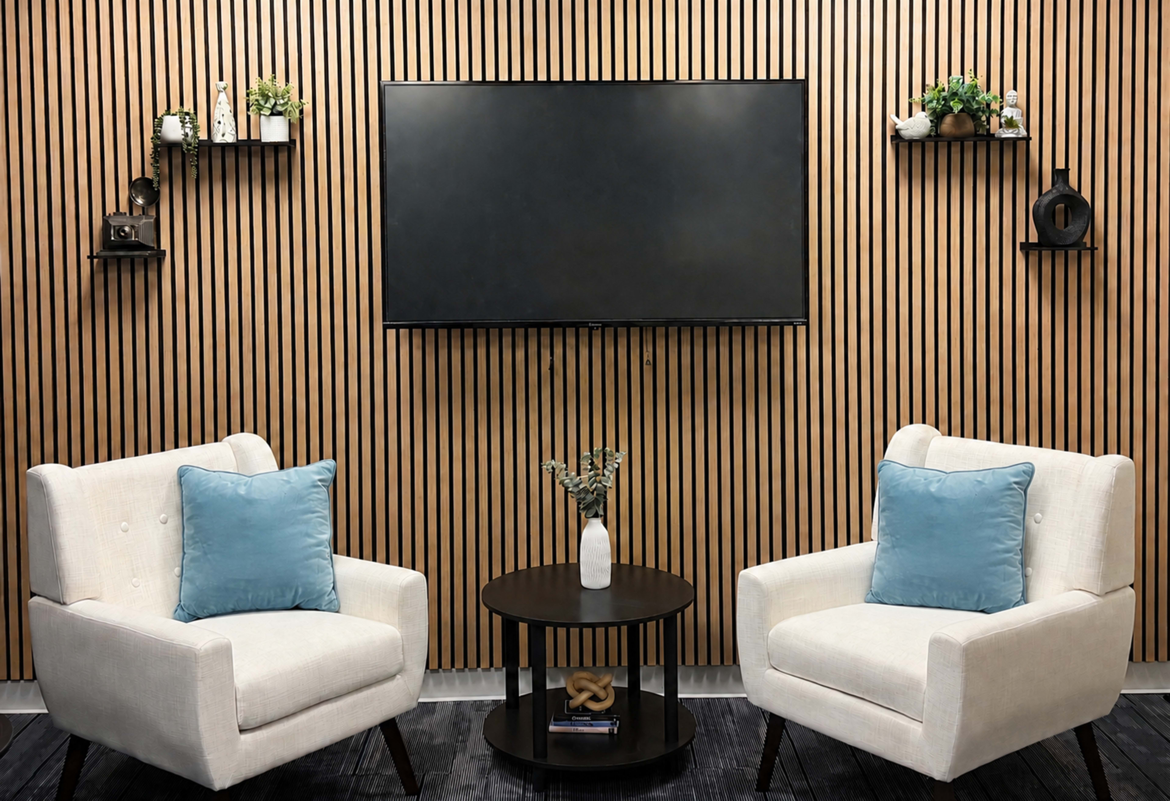 The image shows a modern seating area with two white armchairs featuring blue cushions, positioned around a small circular table, against a backdrop of vertical wooden slats with wall-mounted shelves.
