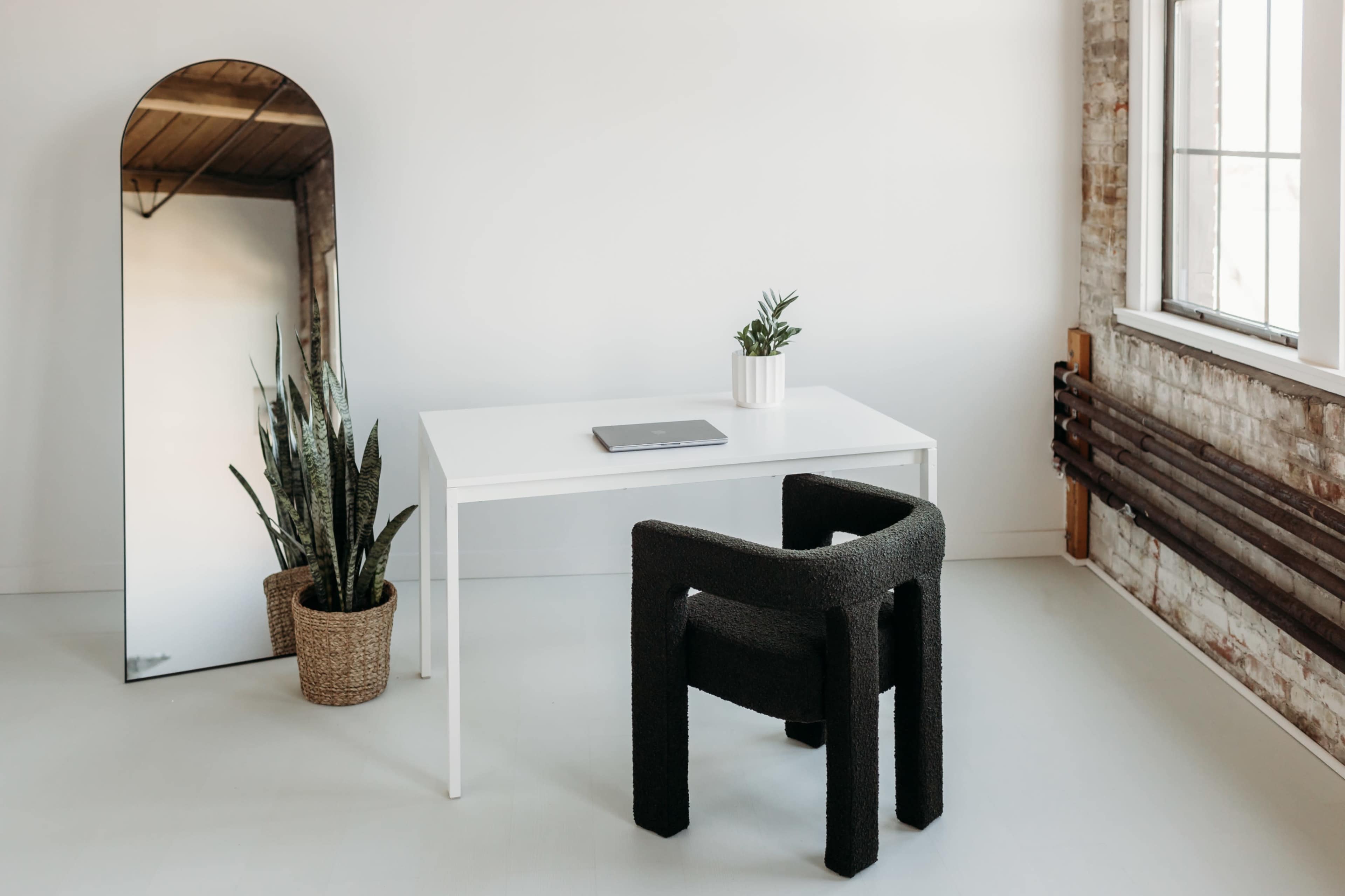 A minimalist workspace features a white table, a black chair, a potted plant, and a large mirror reflecting the room.