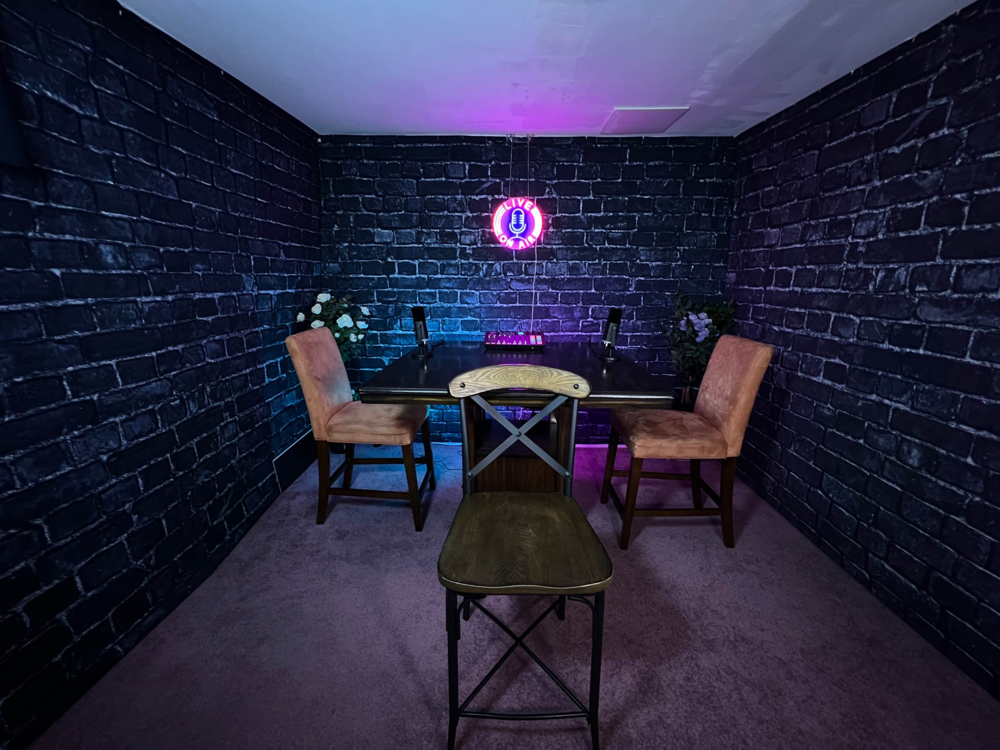 A dimly lit room with a table and chairs against a dark brick wall, featuring a neon sign in the background and floral arrangements on the table.