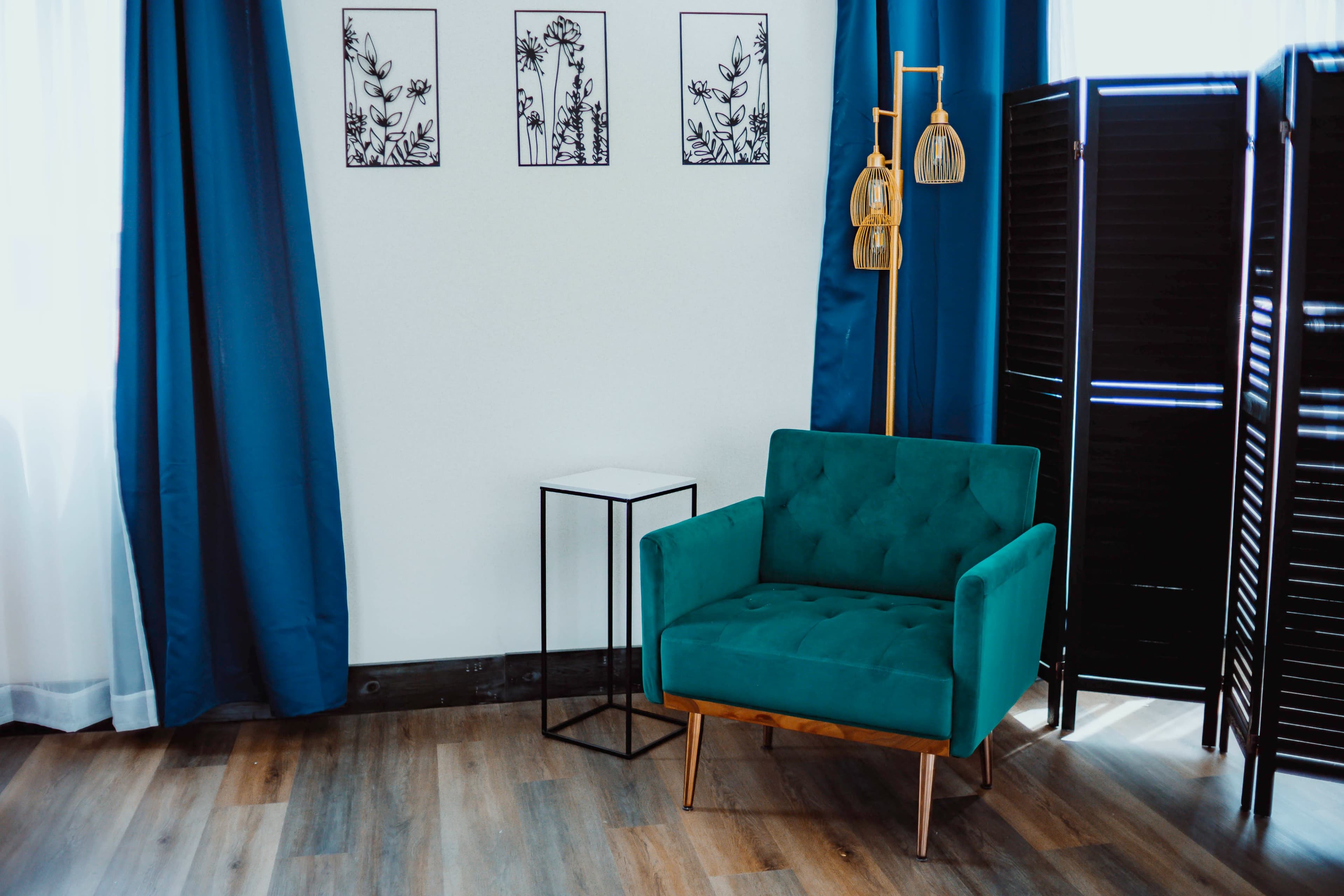 The image shows a cozy living space featuring a teal armchair, a modern side table, decorative wall art, and a folding screen beside blue curtains.