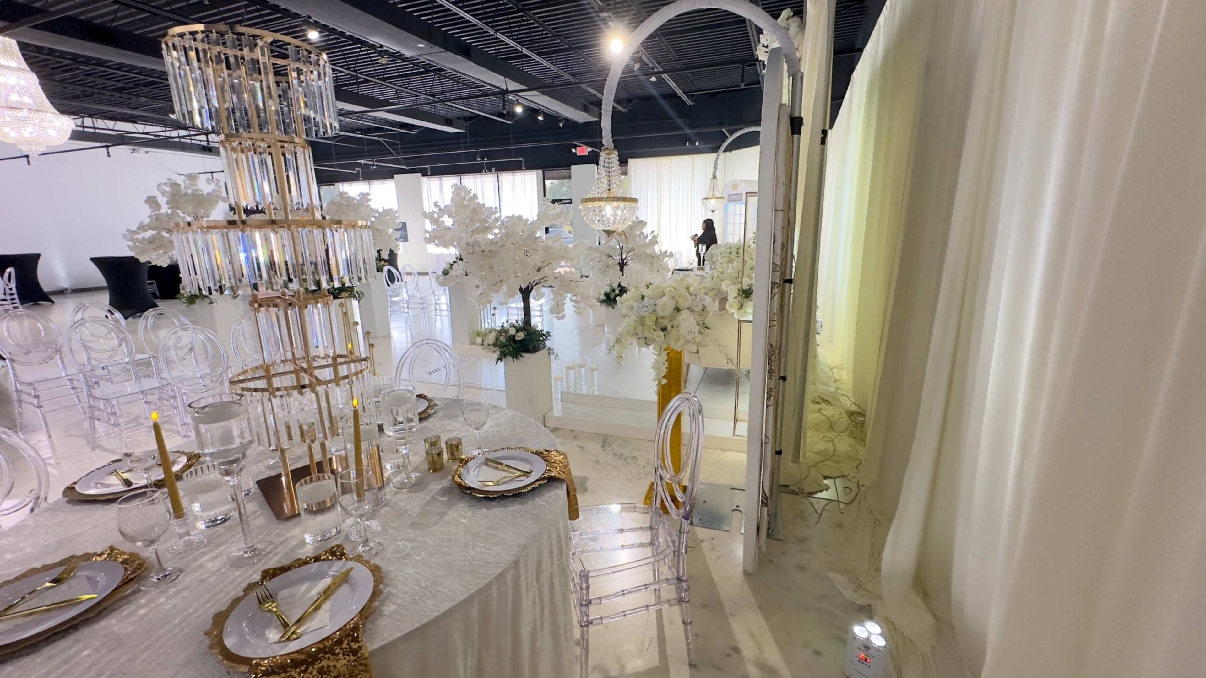 The image shows a elegantly decorated banquet setting with a round table set with fine china and gold accents, surrounded by transparent chairs, and adorned with white floral arrangements in a bright venue.