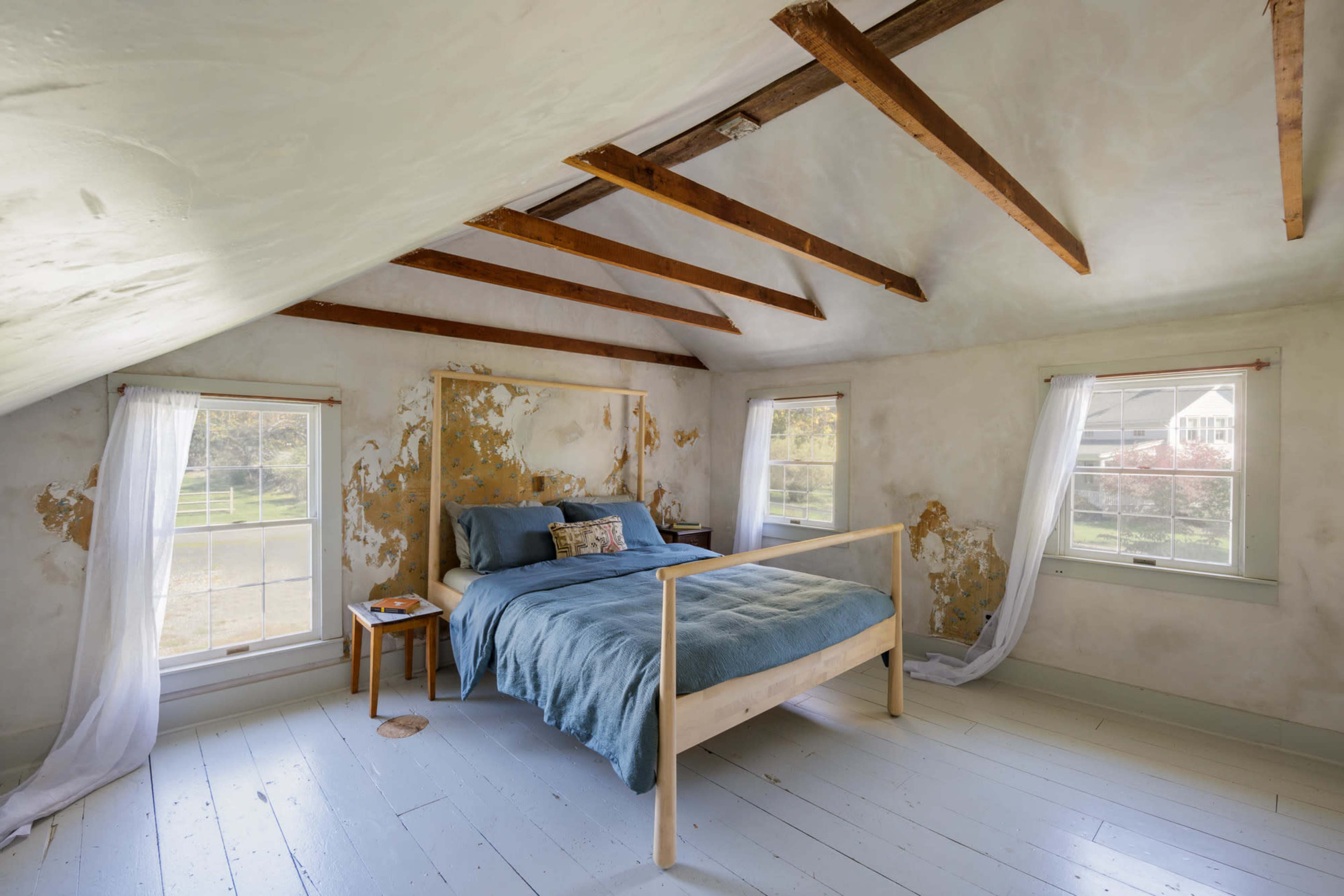 A bedroom features a bed with a blue coverlet, wooden beams overhead, and walls showing signs of wear.