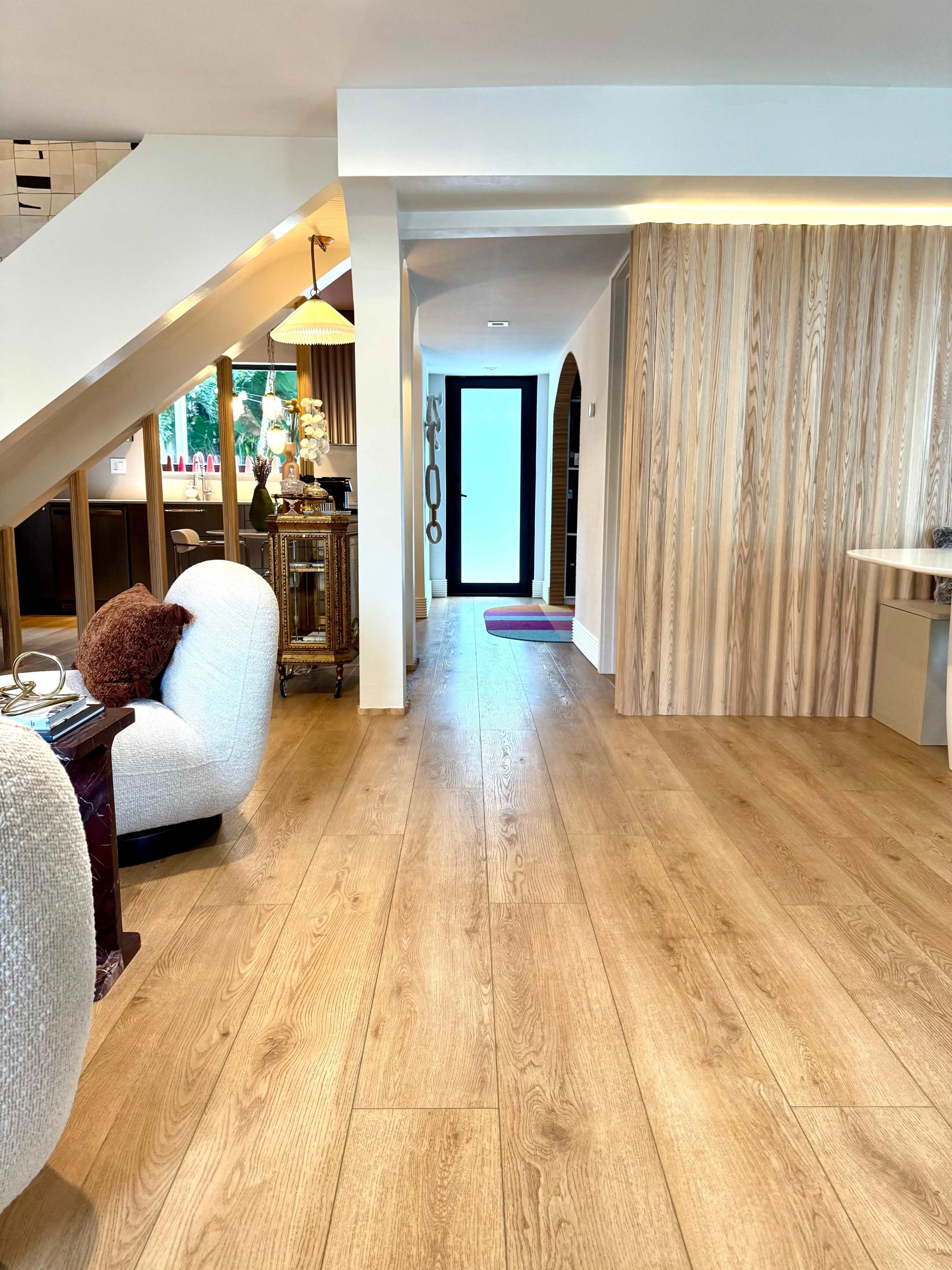 The image shows a modern interior space featuring wooden flooring, a staircase, and a well-lit hallway leading to a door with minimalist decor on both sides.