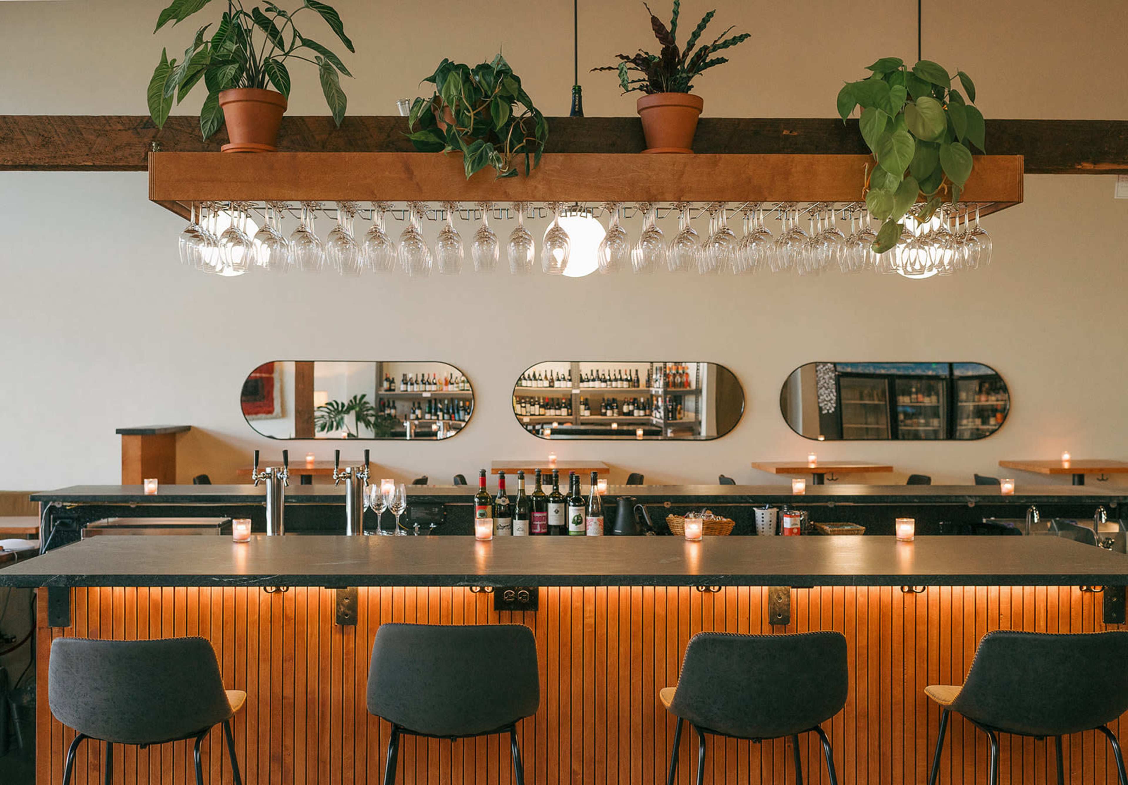 The image shows a modern bar area with a long counter, overhead glassware, and wall mirrors, surrounded by plants and softly lit candles.