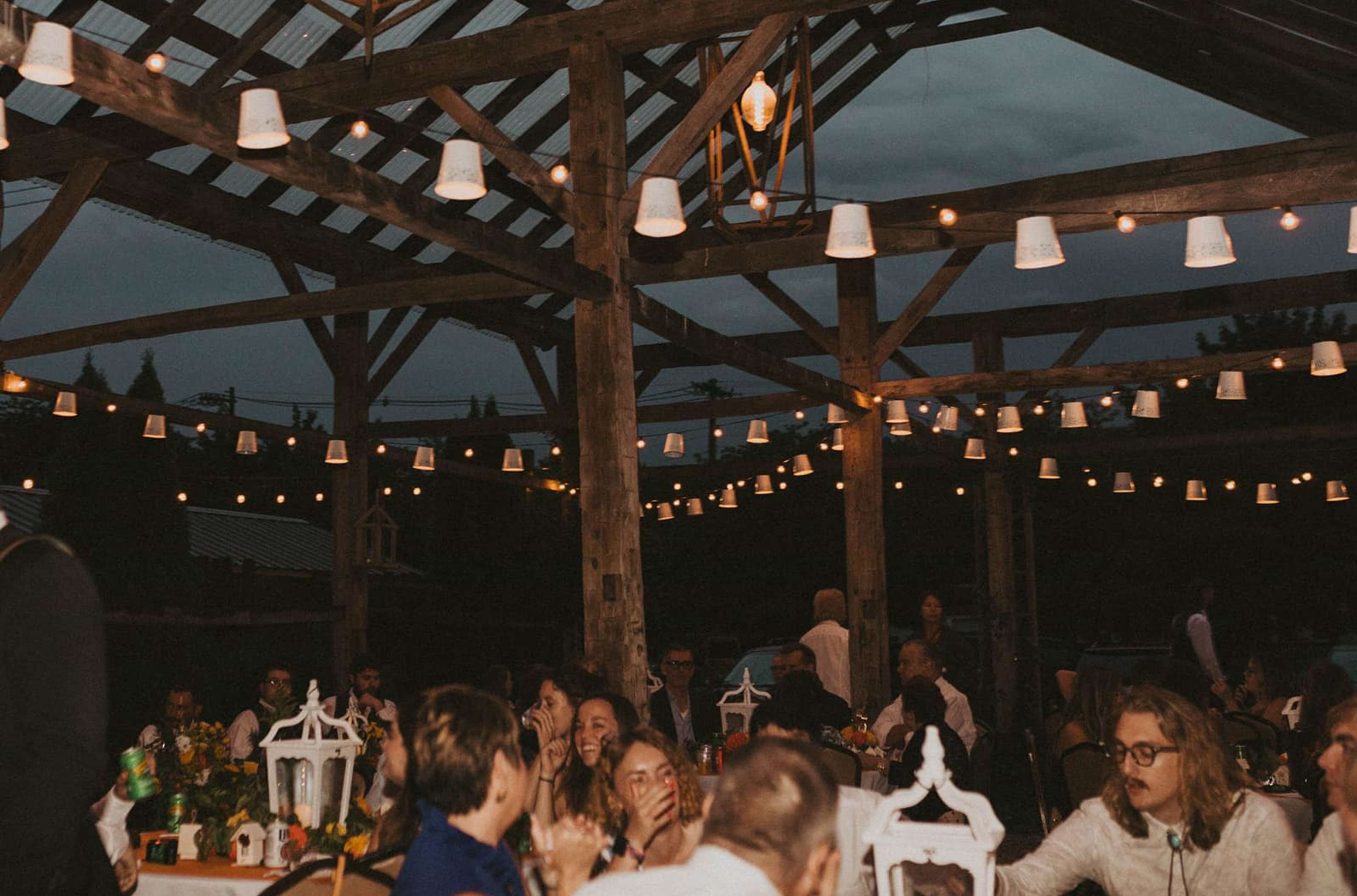 A lively outdoor gathering is set under a wooden structure adorned with string lights, with guests seated at tables enjoying the evening.