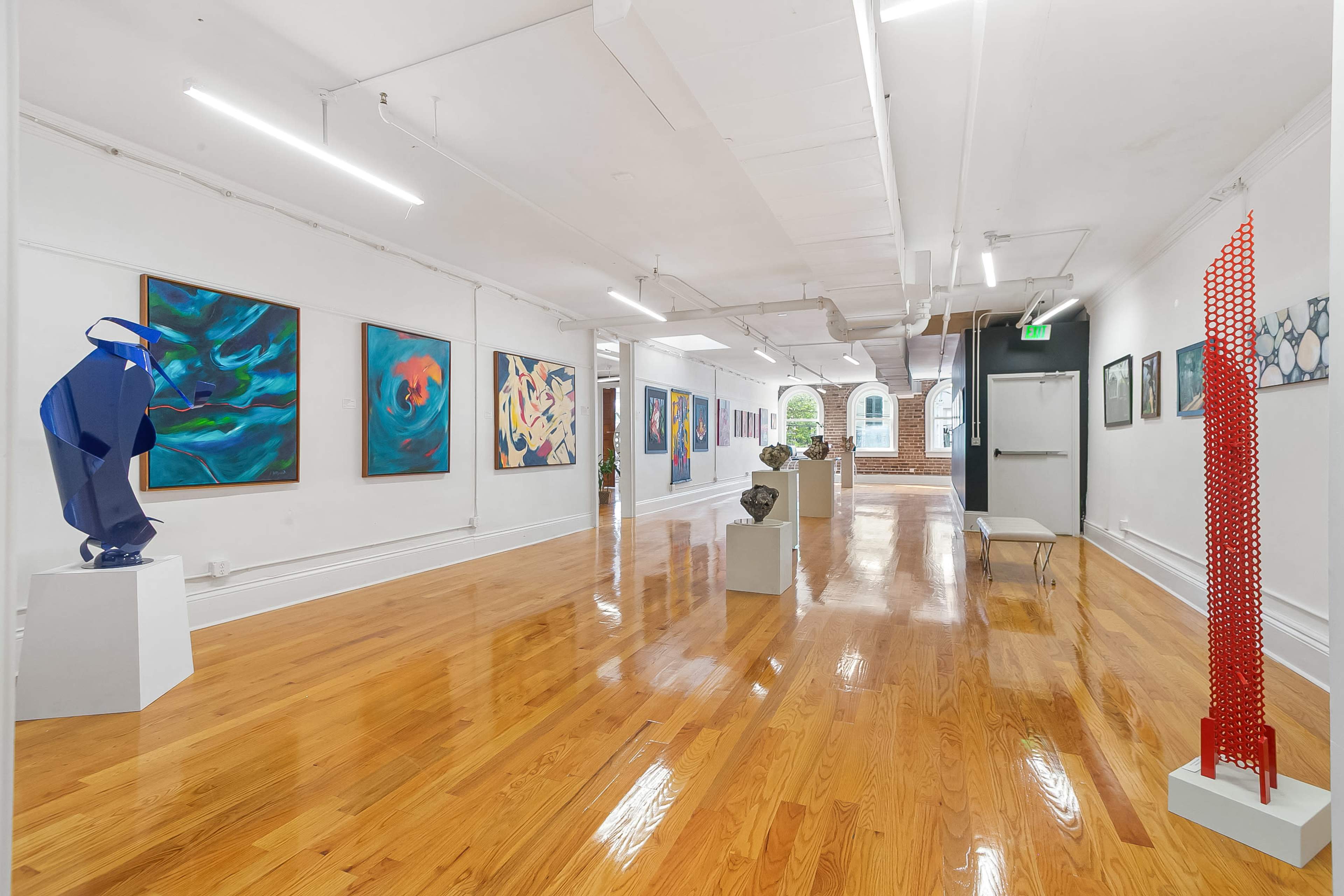 The image depicts a spacious art gallery featuring colorful abstract paintings on the walls and various sculptures displayed on pedestals along a polished wooden floor.