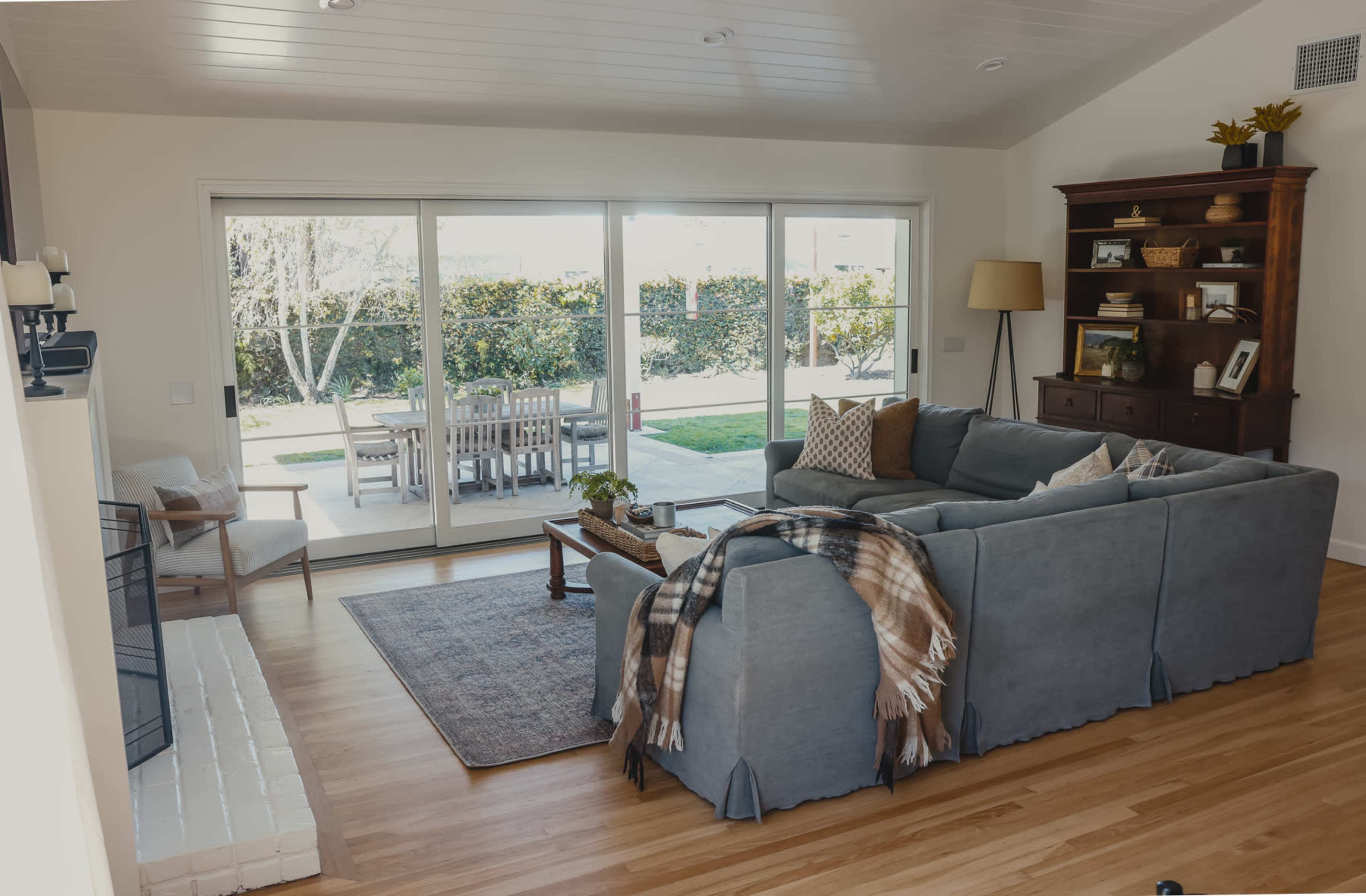 A modern living room features a spacious gray sectional couch, a wooden coffee table, and a bookshelf, with large glass doors opening to a backyard patio.