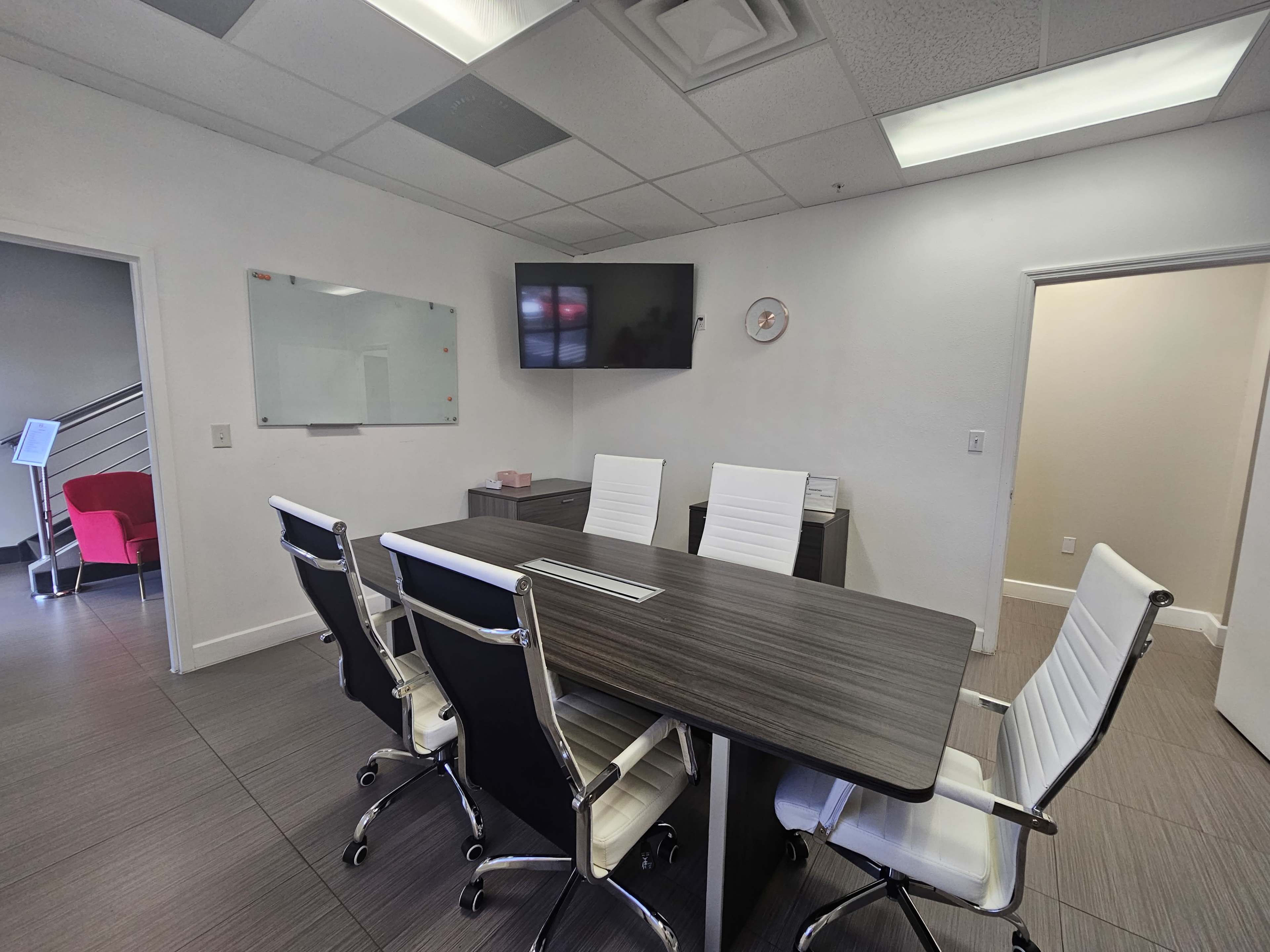 The image shows a modern conference room with a long wooden table, six white ergonomic chairs, a wall-mounted television, and a whiteboard.