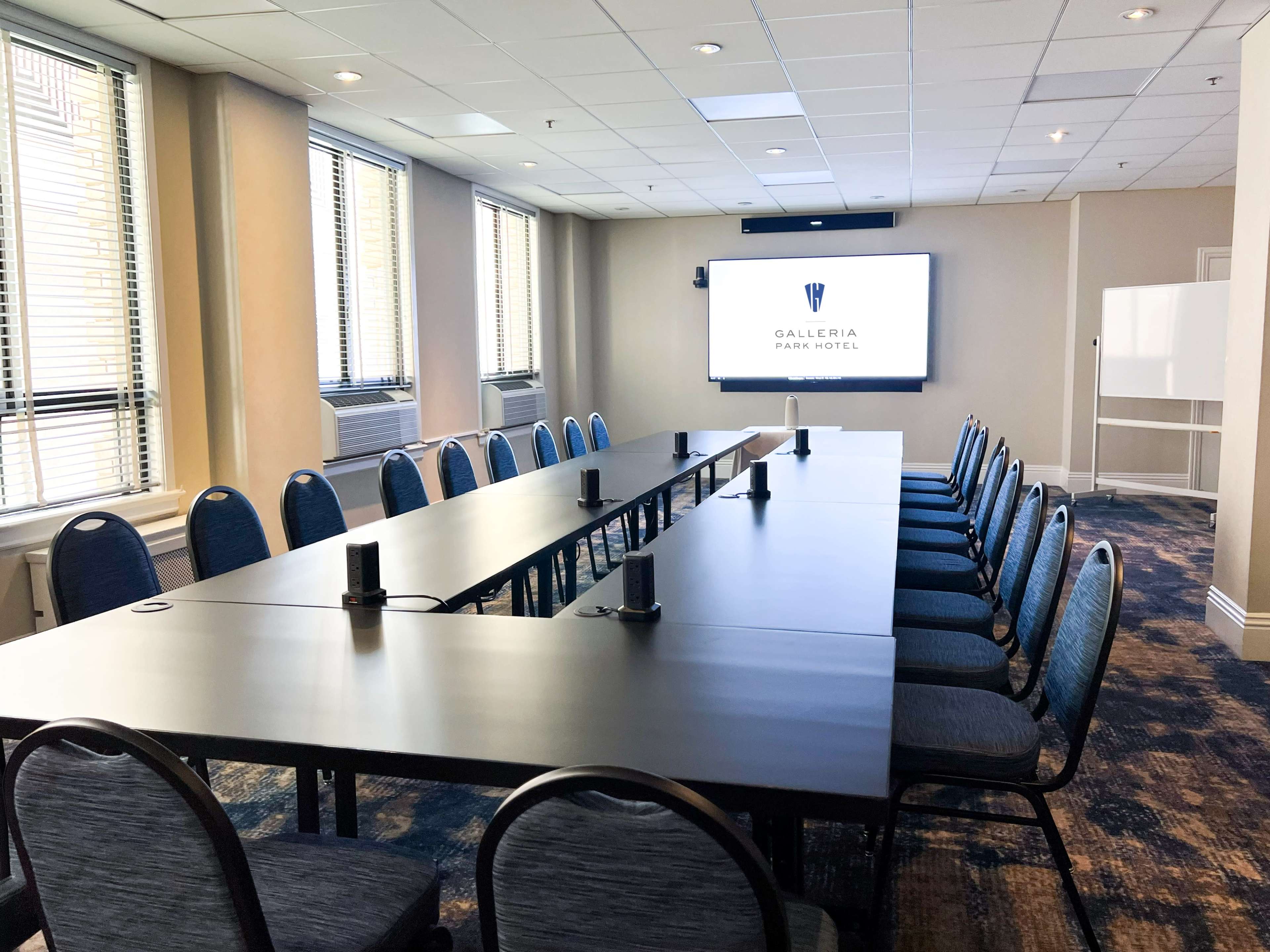 A meeting room with a long table and chairs is set up for a presentation, featuring a large screen displaying the logo of the Galleria Park Hotel.