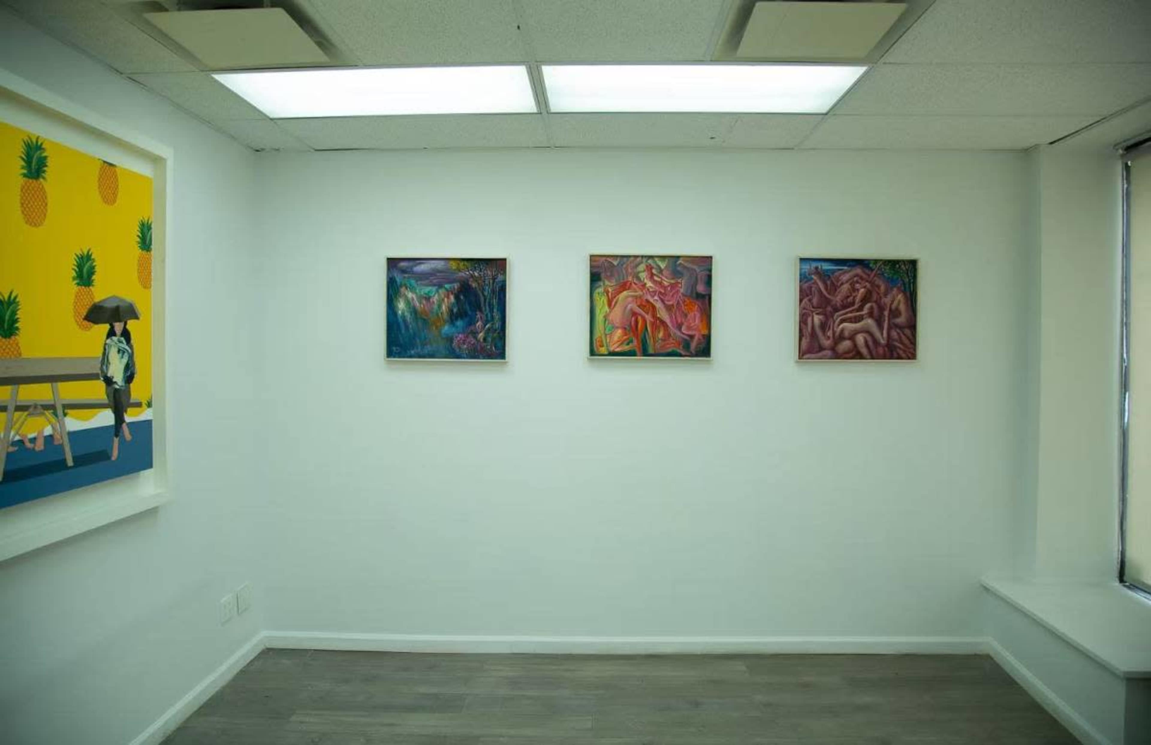 The image shows an art gallery with four framed paintings displayed on white walls, featuring varying abstract styles, alongside a light-colored wall with a pineapple-themed artwork.