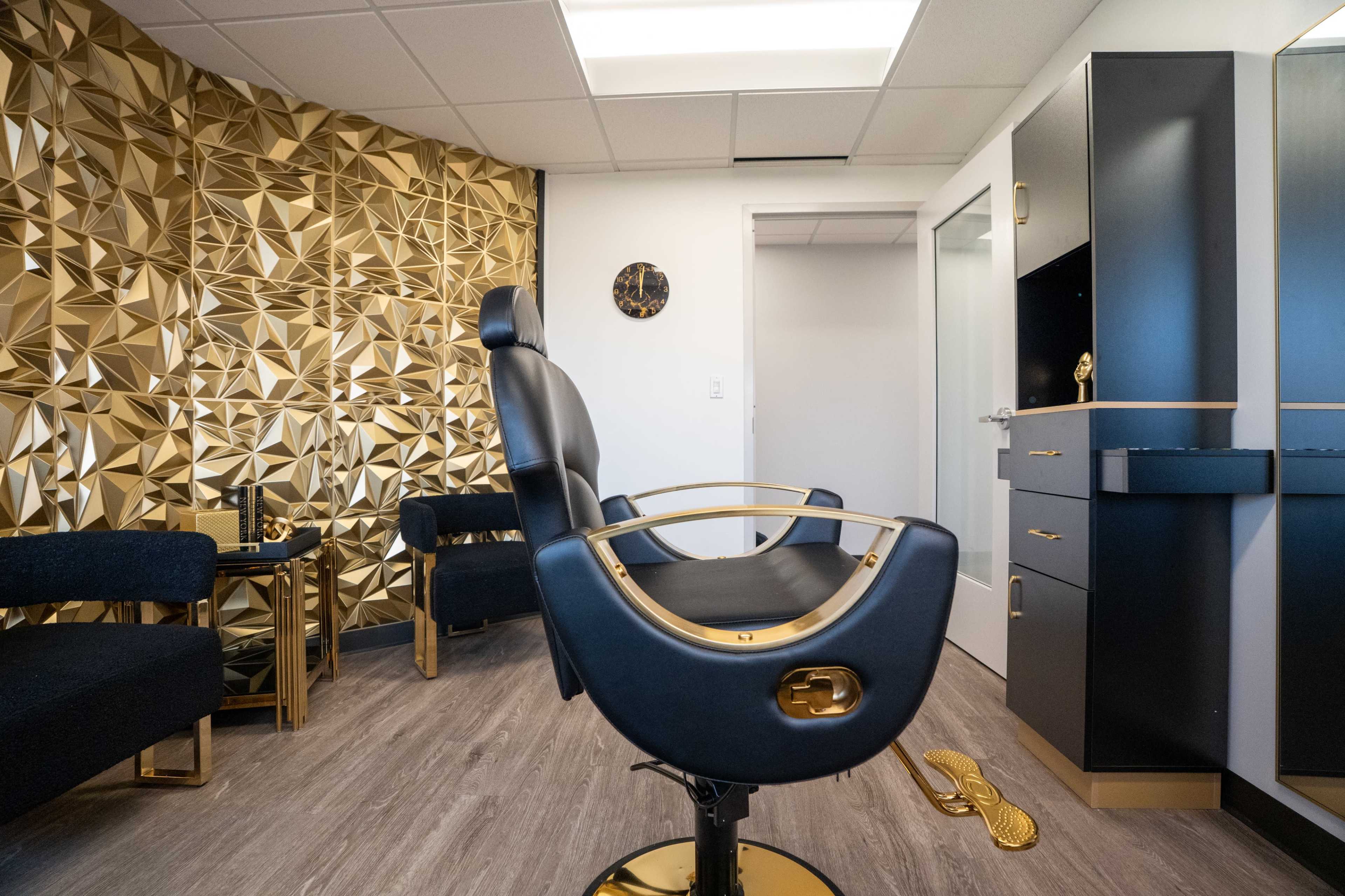 The image shows a modern salon room featuring a black and gold barber chair, geometric gold wall decor, and a minimalist vanity area.