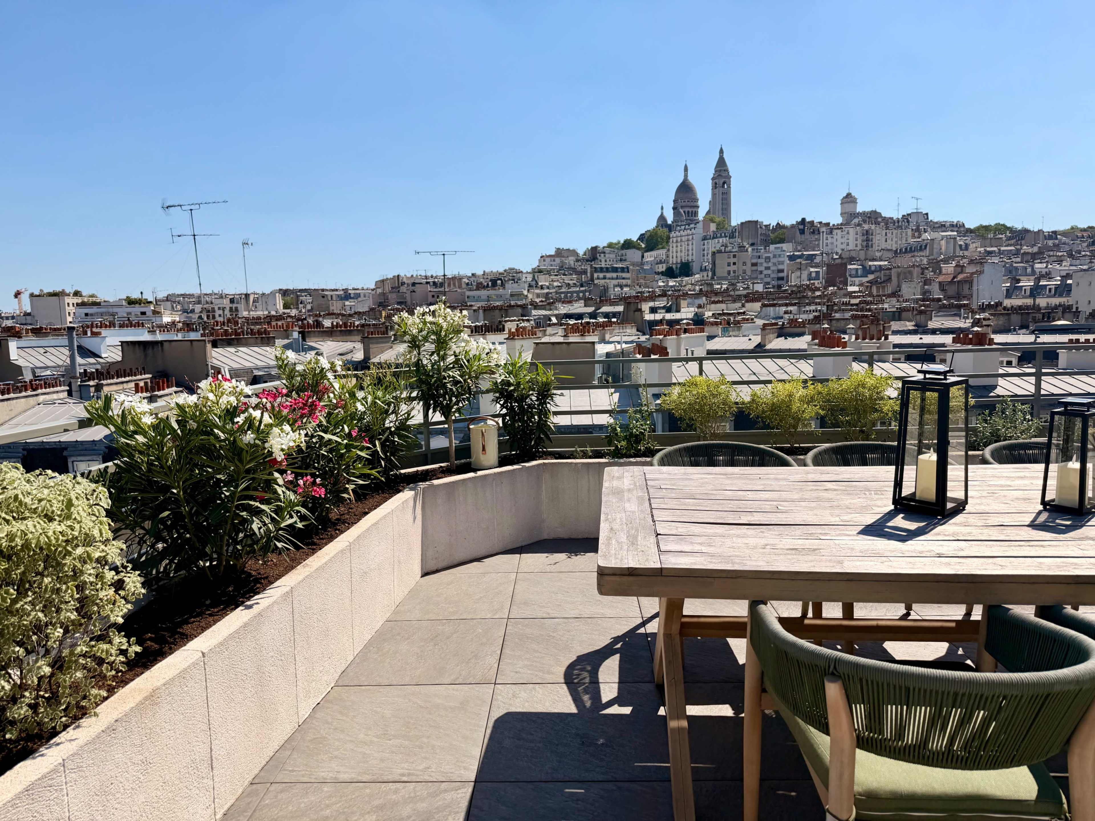A rooftop terrace features a wooden table and chairs with colorful flowers and plants, overlooking a cityscape with distant hills and buildings including a church.