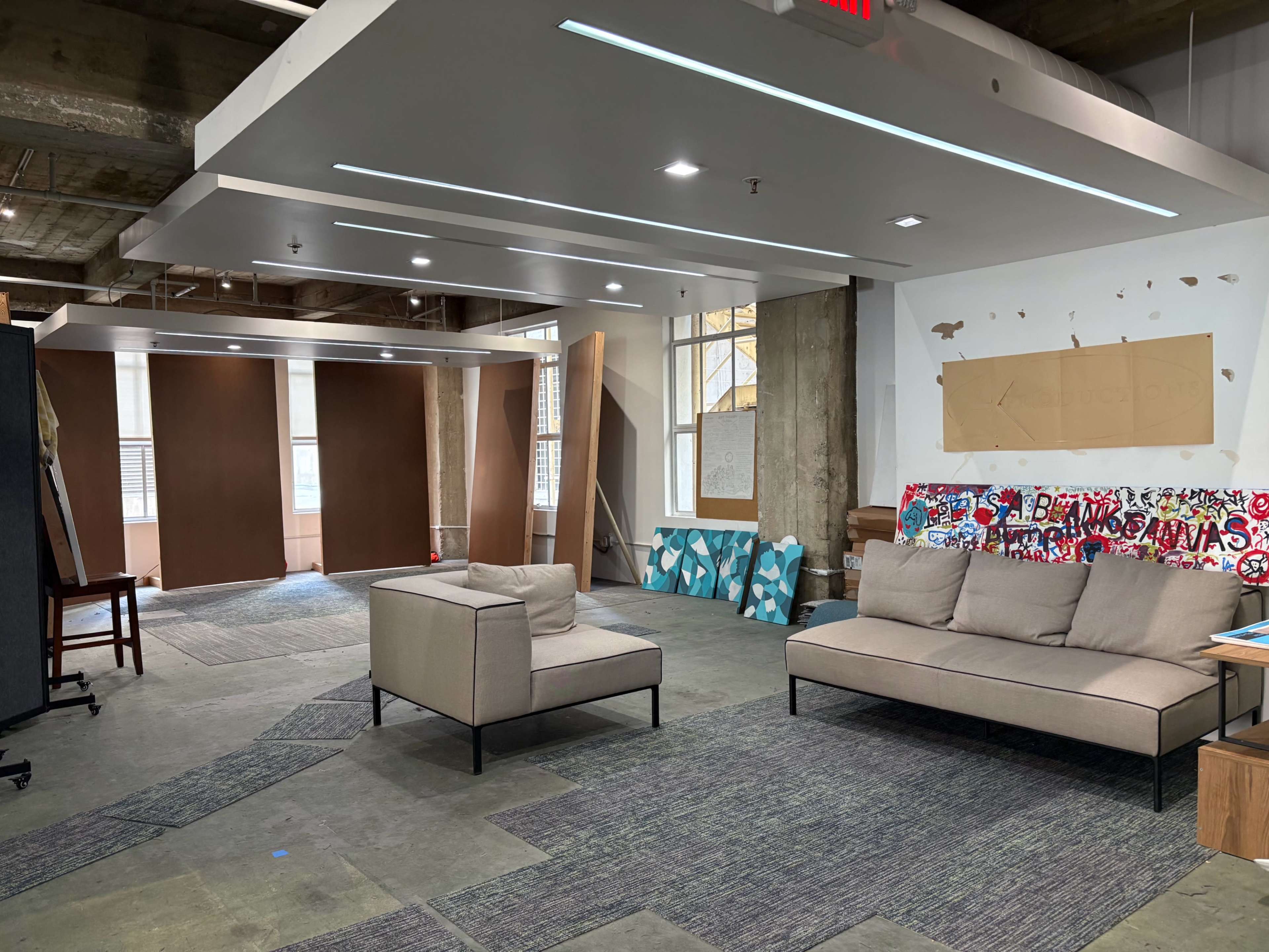 The image shows a spacious interior room with modern furniture, including a light-colored sofa and a chair, against a backdrop of exposed concrete walls and a large window.