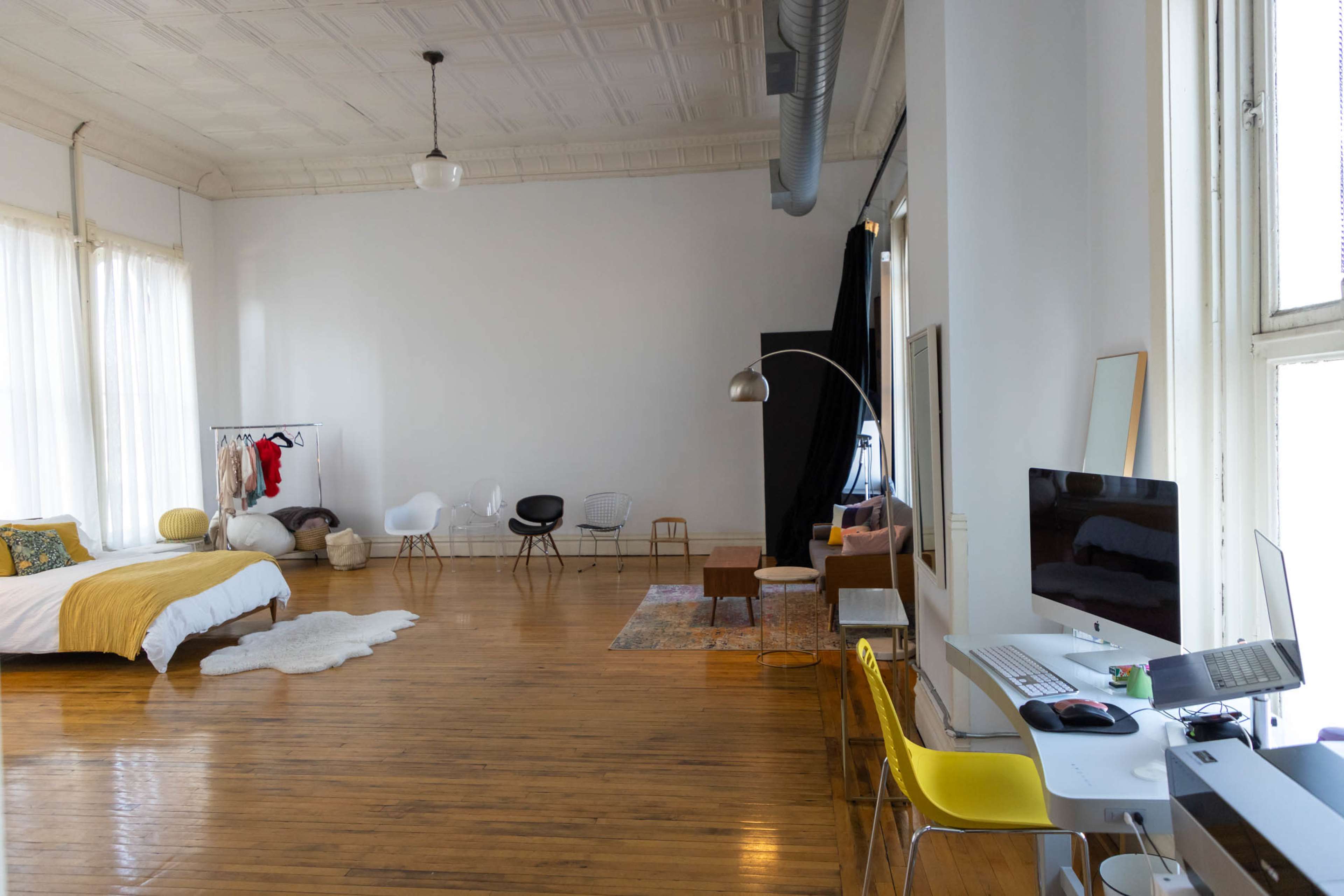 The image shows a spacious, well-lit room featuring a bed with yellow bedding, a desk with a computer, various seating options, and a clothing rack against a white wall.