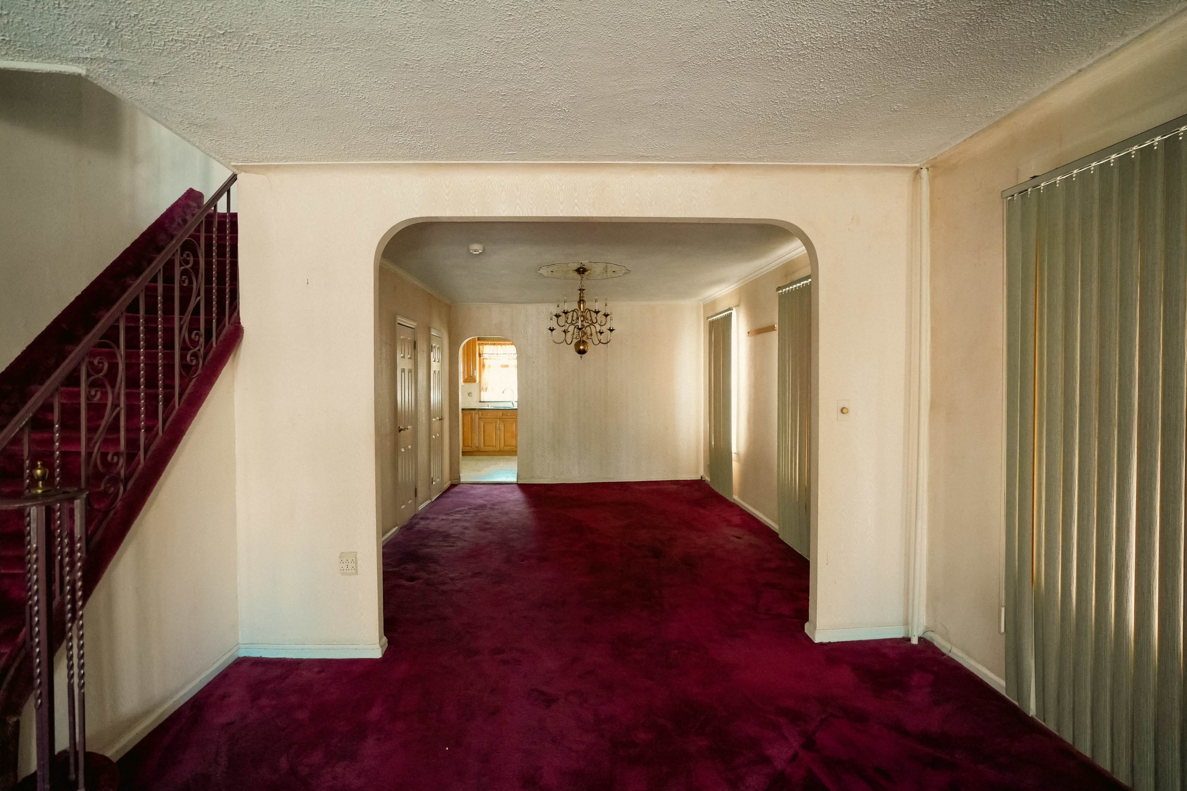 The image shows a spacious living area with pink carpeting, a chandelier hanging from the ceiling, and an archway leading to a kitchen area.