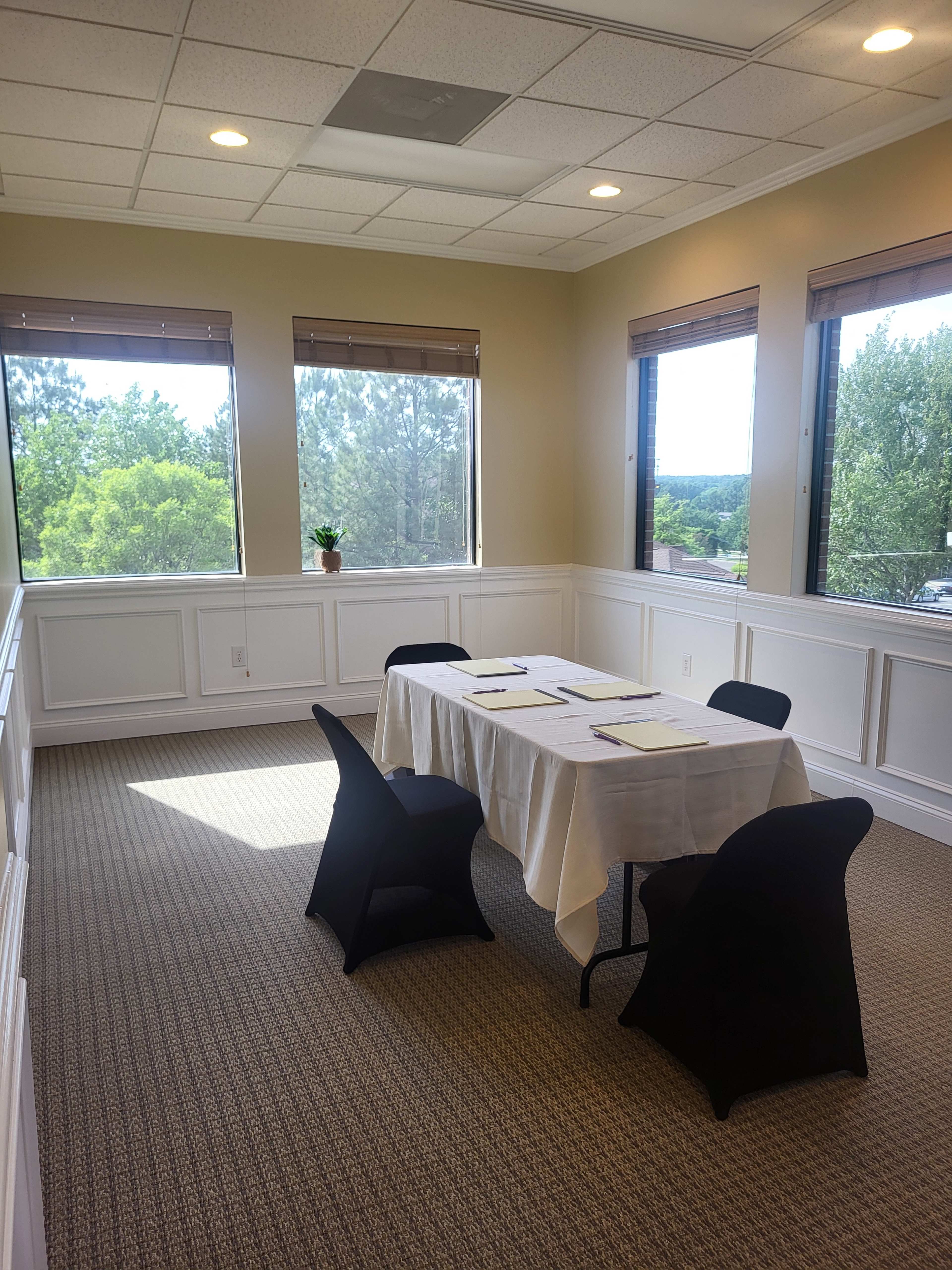 A small conference room features a table set for a meeting, surrounded by four black chairs, and three large windows provide natural light and a view of greenery outside.