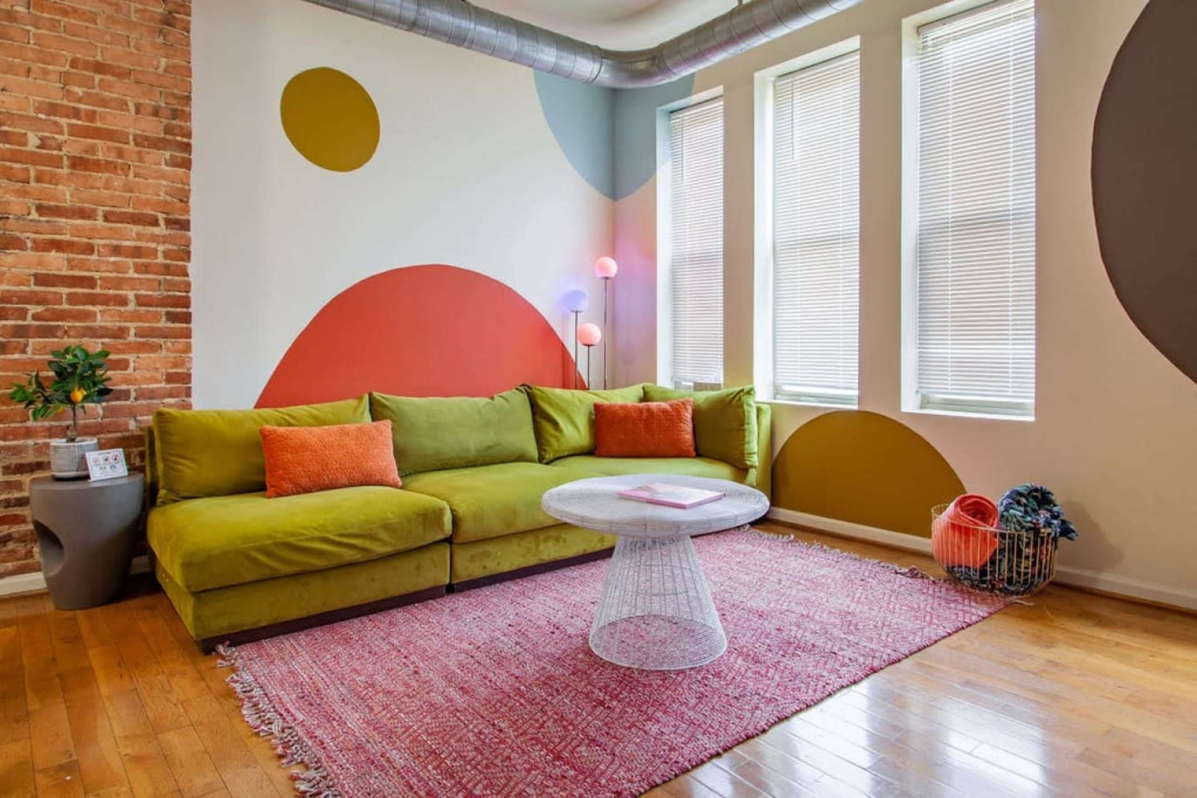 A vibrant living room features a green sectional sofa, a round glass coffee table, and colorful wall accents, all set against exposed brick and large windows.